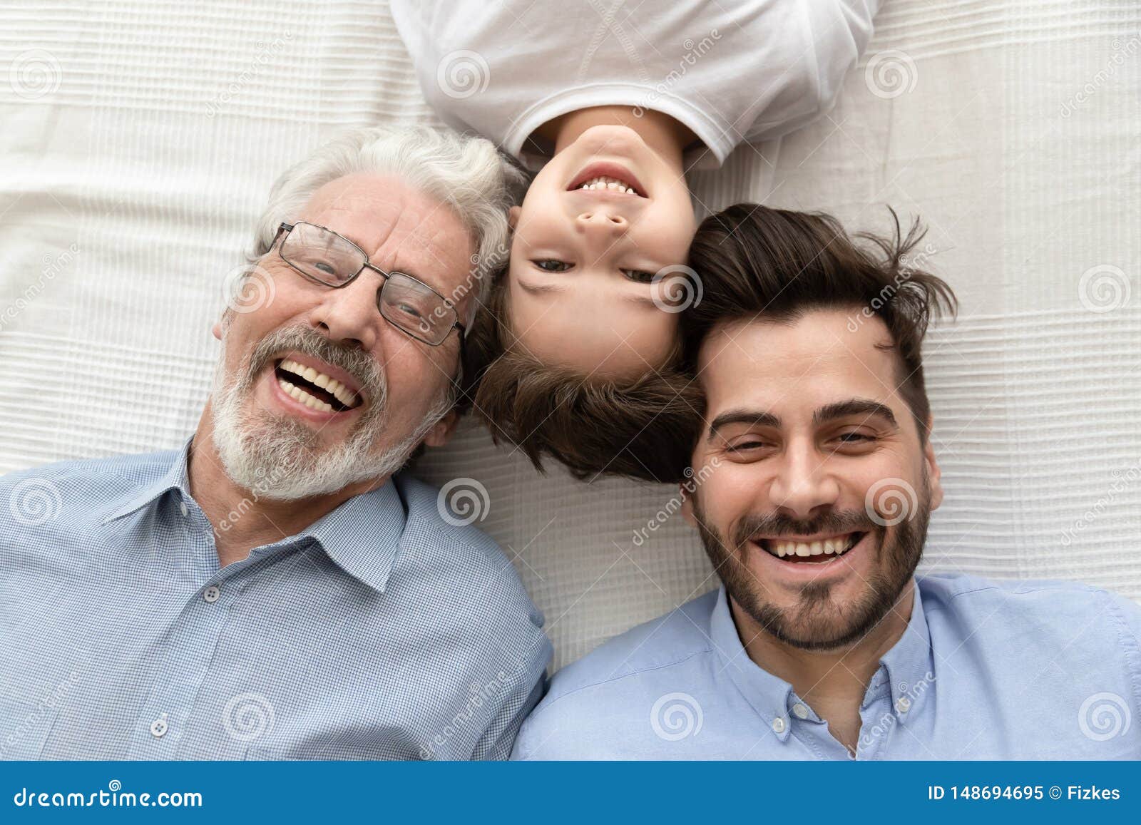 Opini?n superior tres generaciones felices de sonrisa de los hombres. Top view of happy three generations of men lying on bed looking at camera making photo together, portrait of smiling little son, father and grandfather posing for picture laughing relaxing at home