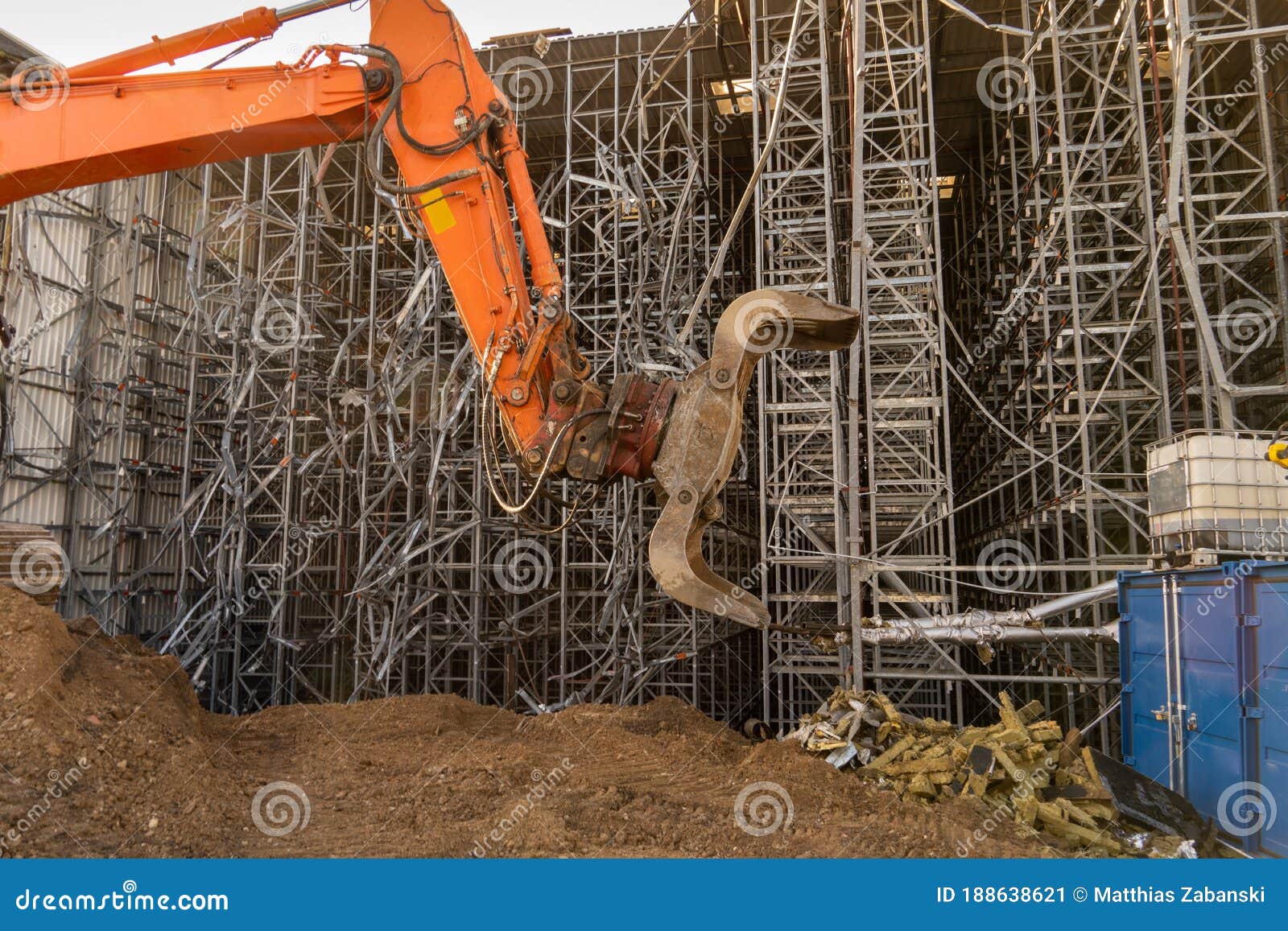orange gripper with excavator bucket in front of a high-bay warehouse that is being torn down