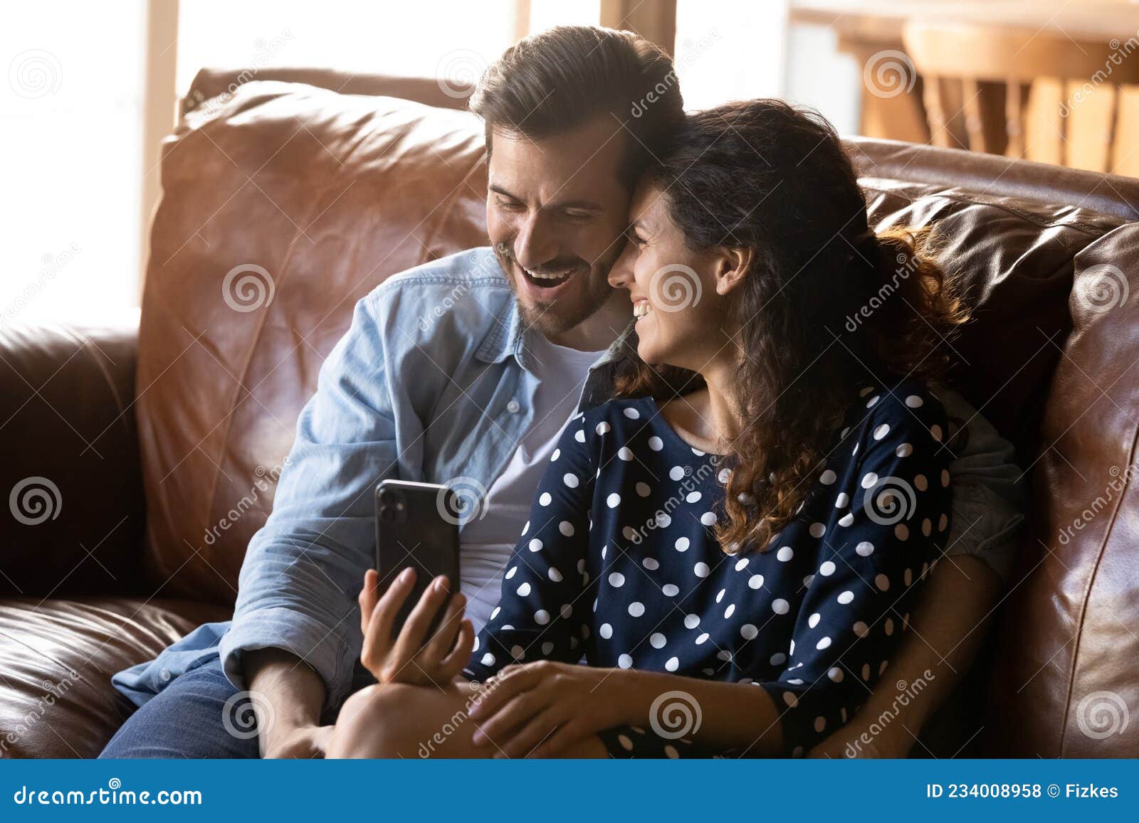 Overjoyed Young Couple Cuddle on Sofa Watch Comedy on Cell Stock Photo