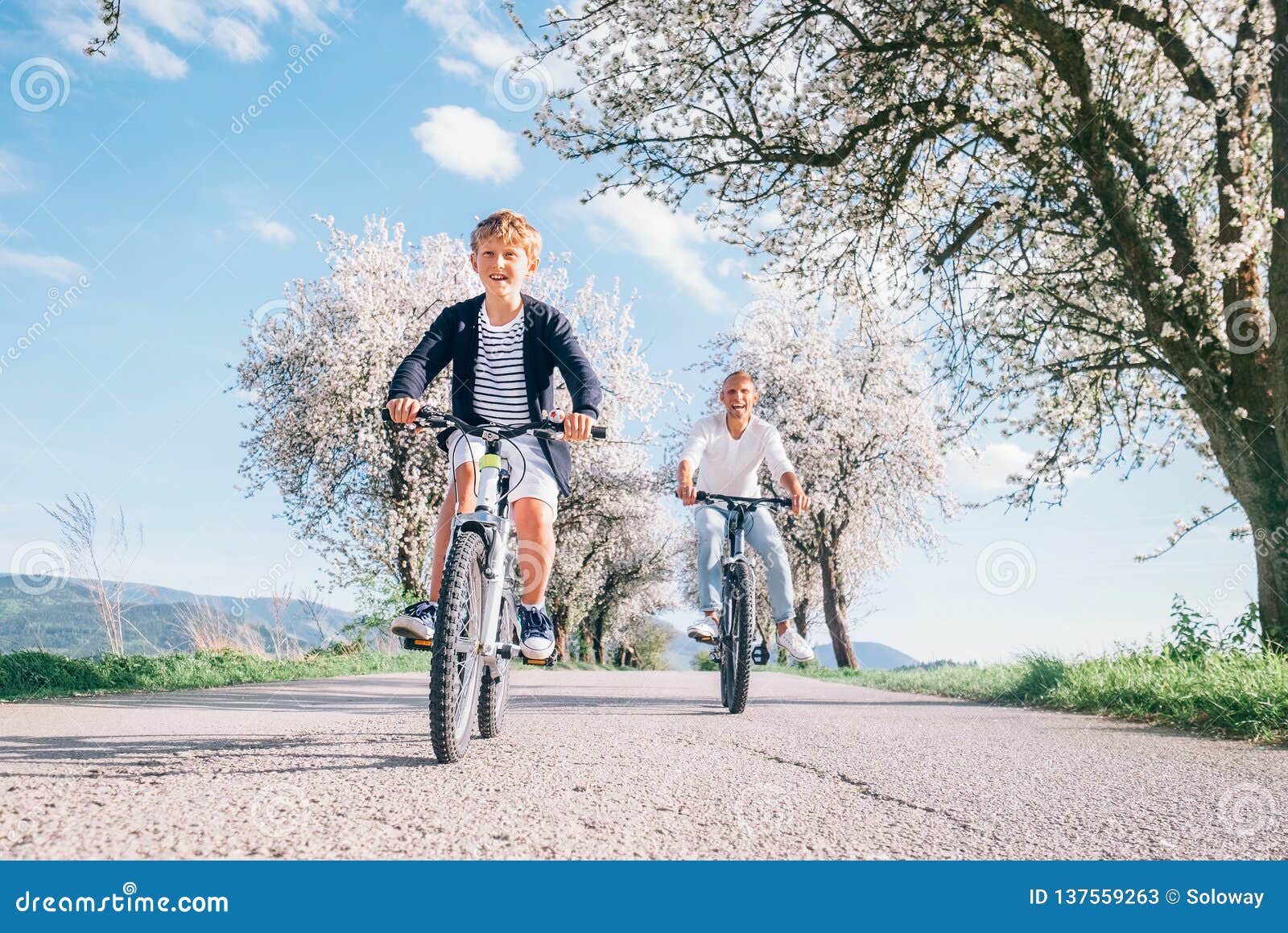 Padre e figlio divertendosi quando guidano le biciclette sulla strada campestre sotto gli alberi del fiore Immagine sportiva sana di concetto di stile di vita
