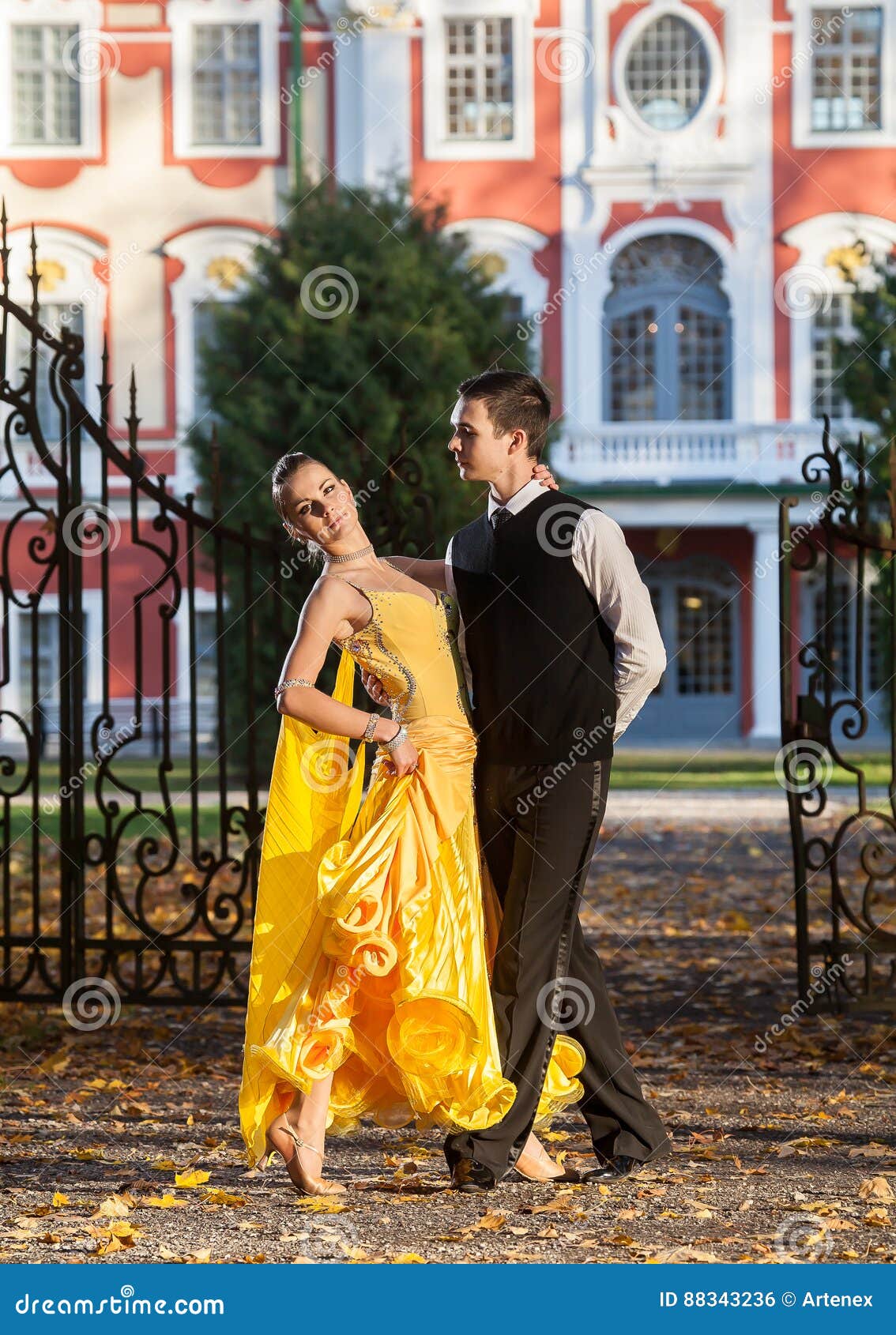 Pair of dancers dancing in the woods. Man with suit, woman in yellow long dress middle of the palace park in autumn. Dry fallen c. Pair of dancers dancing in the palace garden. Man with suit and women in a yellow long dress in the middle of the park in autumn. Dry fallen colored leaves, trees gates and castle in background