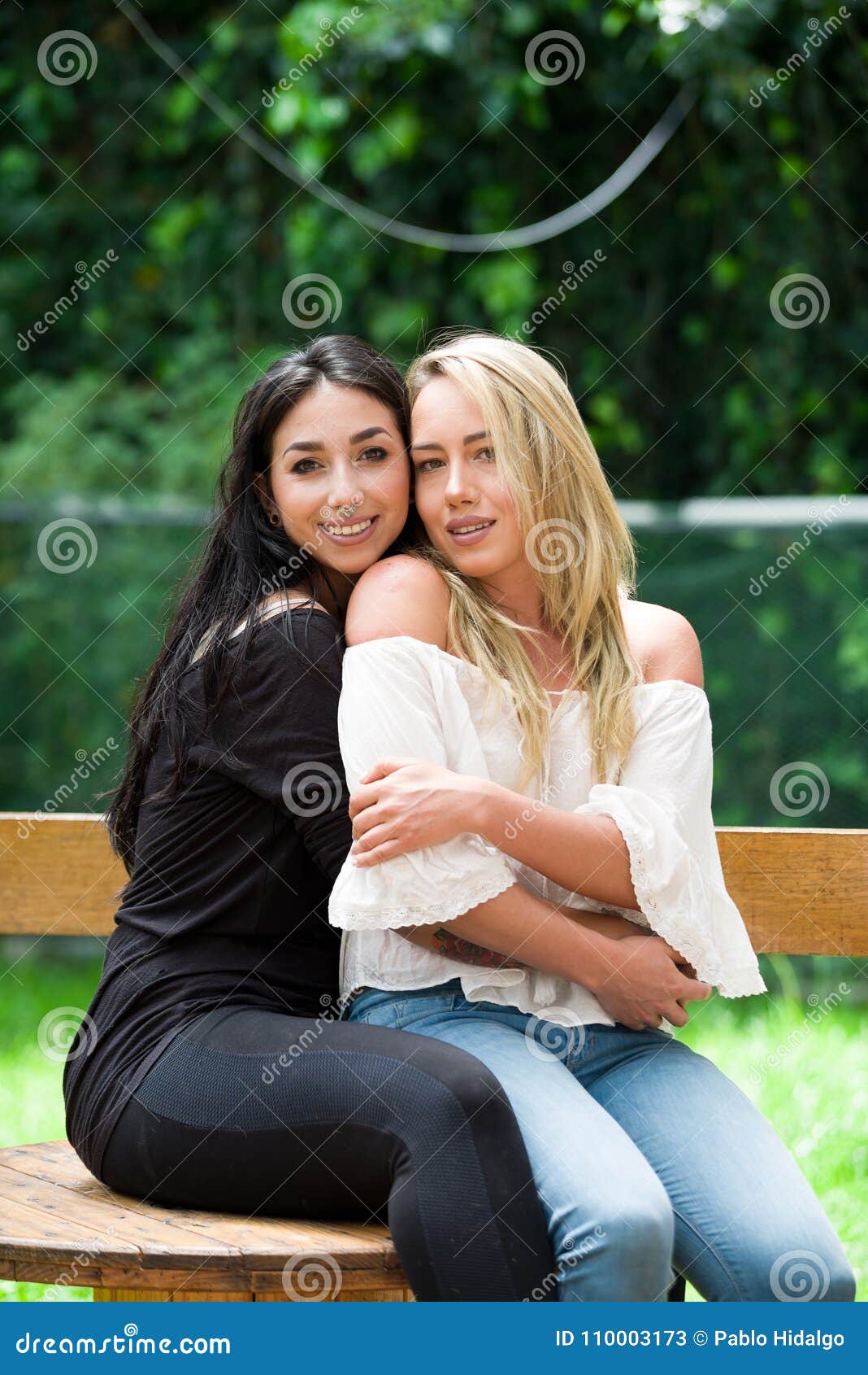 a pair of proud lesbian in outdoors sitting on a wooden table, brunette woman is hugging a blonde woman, in a garden