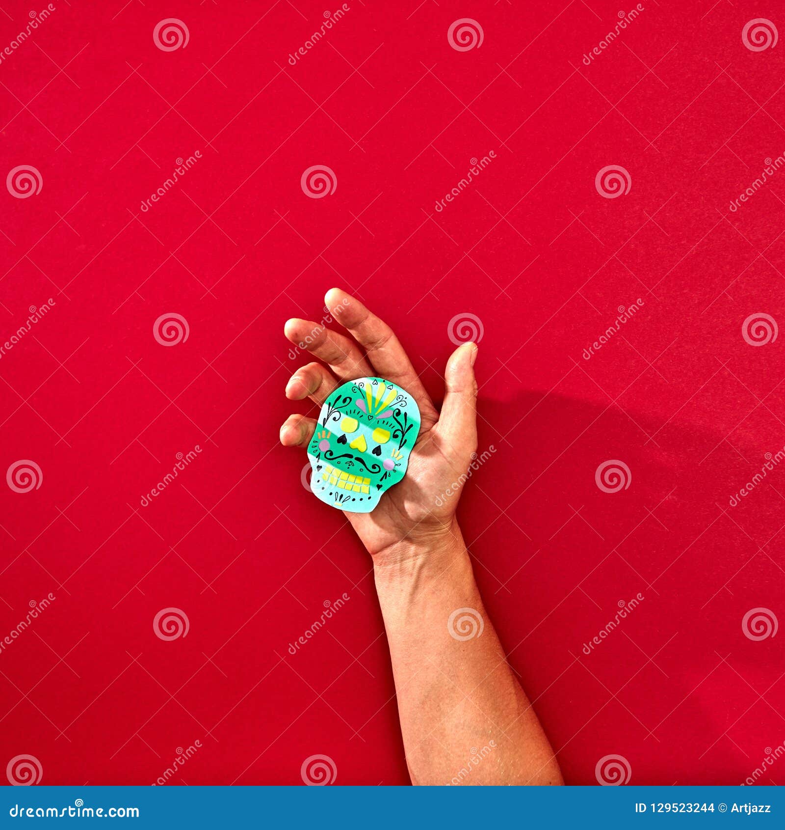 the hand of a man holds a handcraf paper skull calaveras attributes of the mexican holiday calaca on a red background