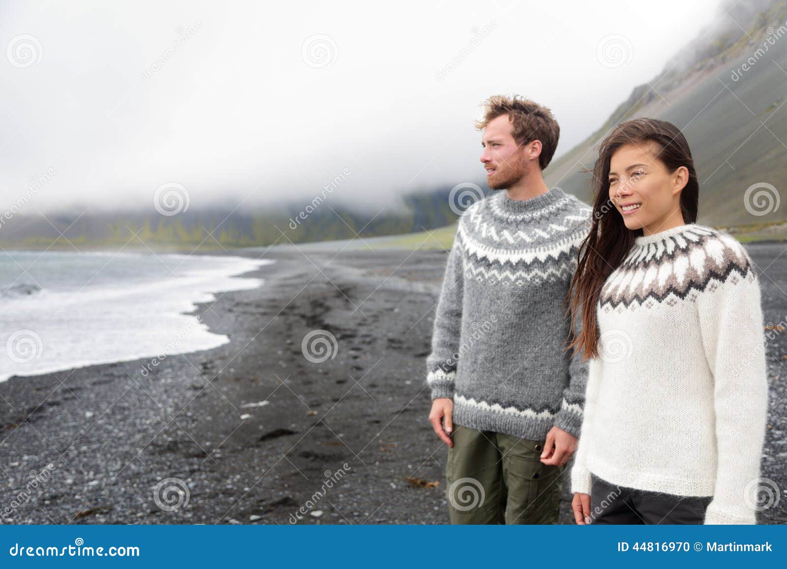 Pares de Islândia que vestem camisetas islandêsas na praia preta da areia A mulher e os homens modelam na camiseta islandêsa típica que olha o oceano que apreciam a ideia da paisagem da natureza no mar do oceano