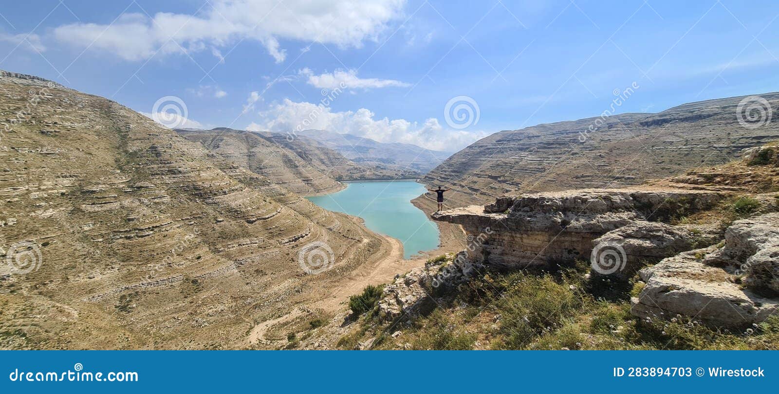 person stands on the edge of a tall cliff overlooking the tranquil lake of faraya