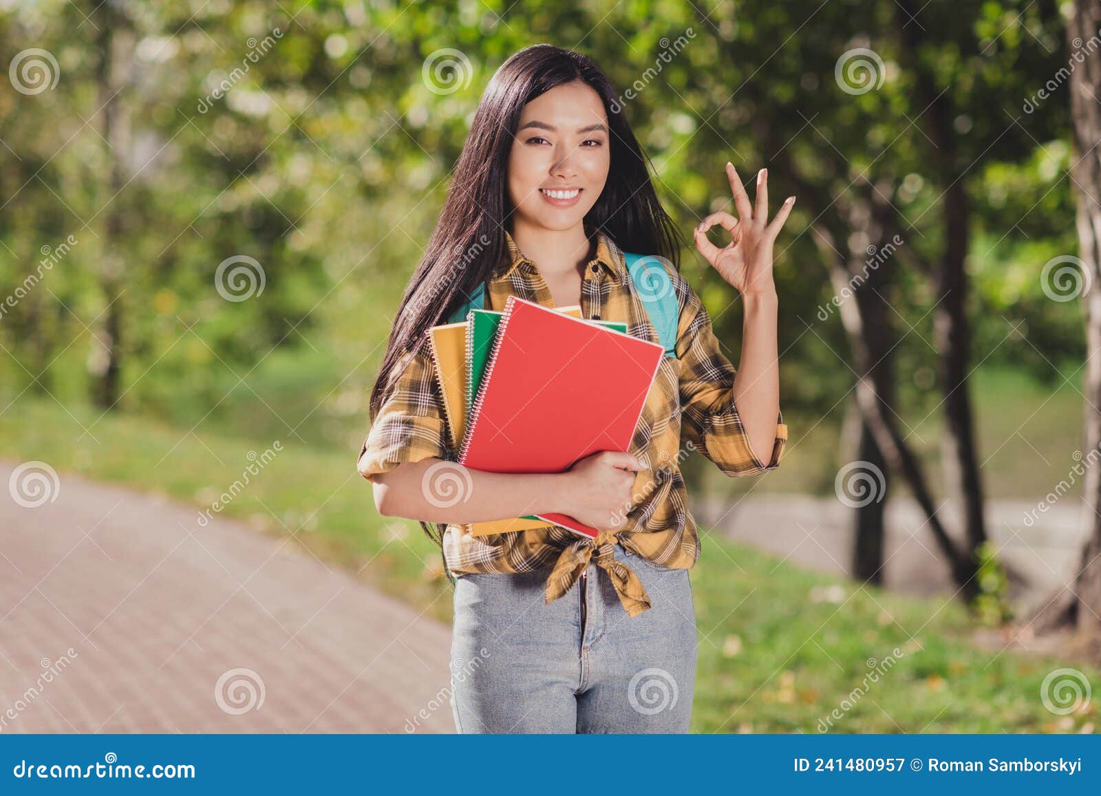photo of adorable funny young lady wear checkered shirt backpack smiling walking holding book showing okey outdoors