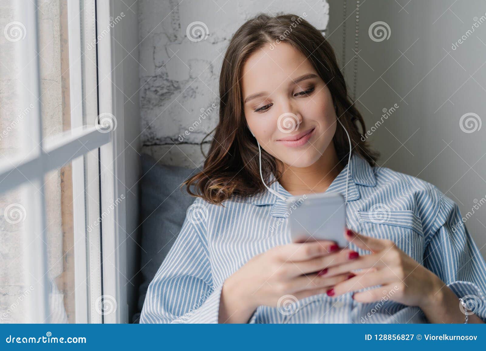 photo of cauasian woman selects favourite song in playlist, uses contemporary cellular and high quality earphones, dressed in casu