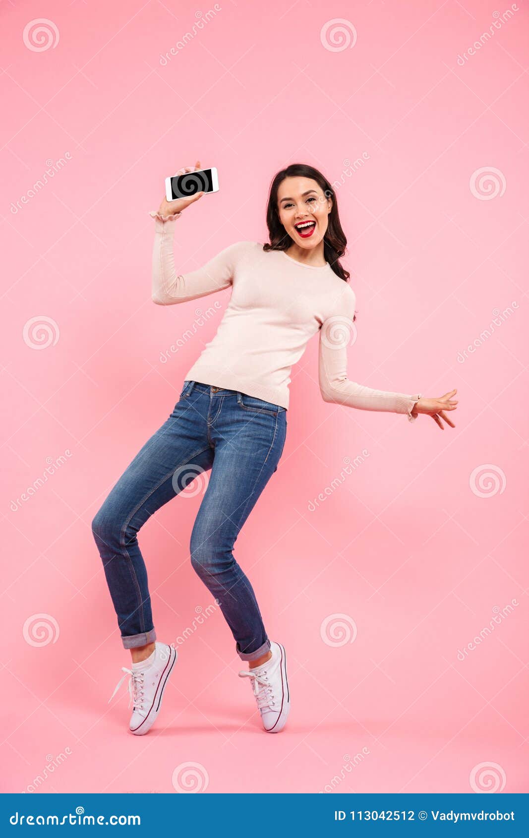 Photo intégrale des jeans de port de la jeune femme 20s démontrant le téléphone portable et exprimant le bonheur d'isolement au-dessus du fond rose