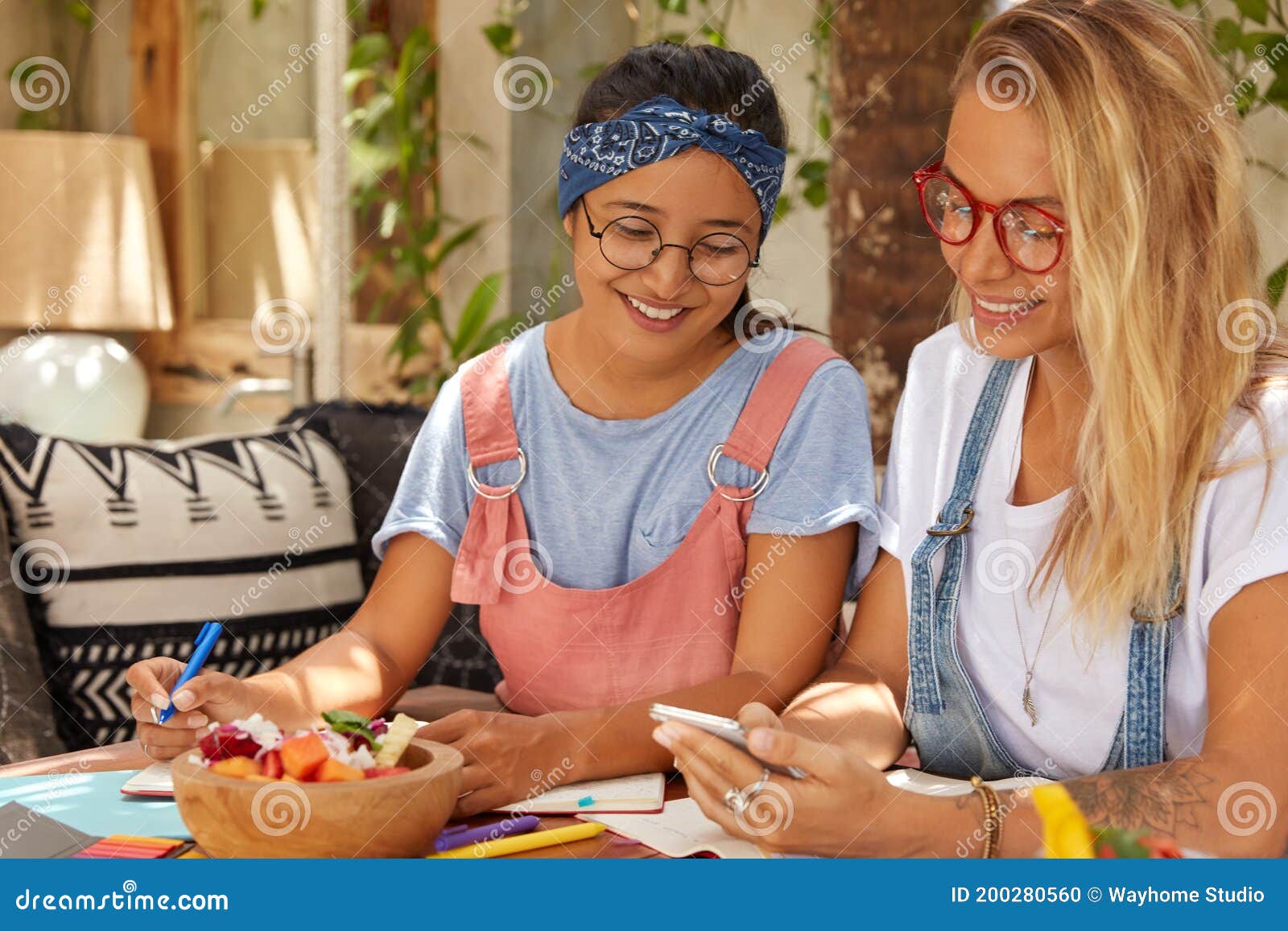 photo of multiethnic girls sit together at cozy interior, use cellulars for reading news and messaging, hold pen, eat