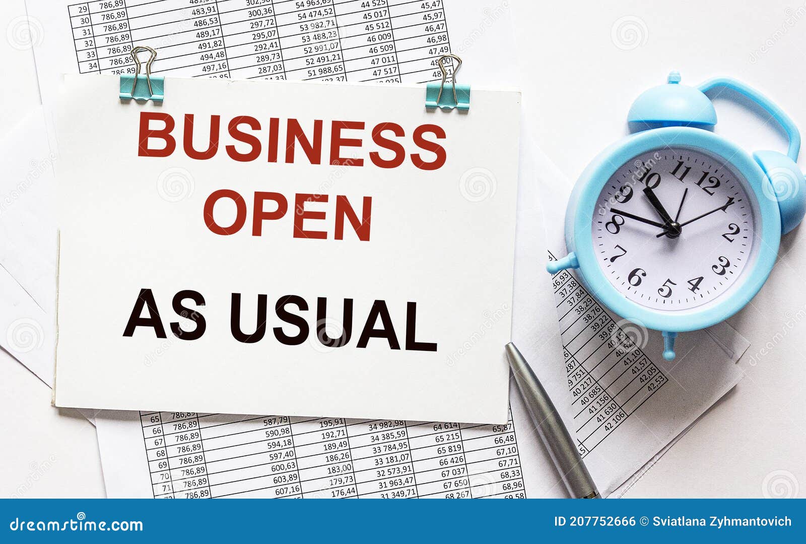 the phrase business as usual printed on a notebook. documents, alarm clock and pen lie on a white background