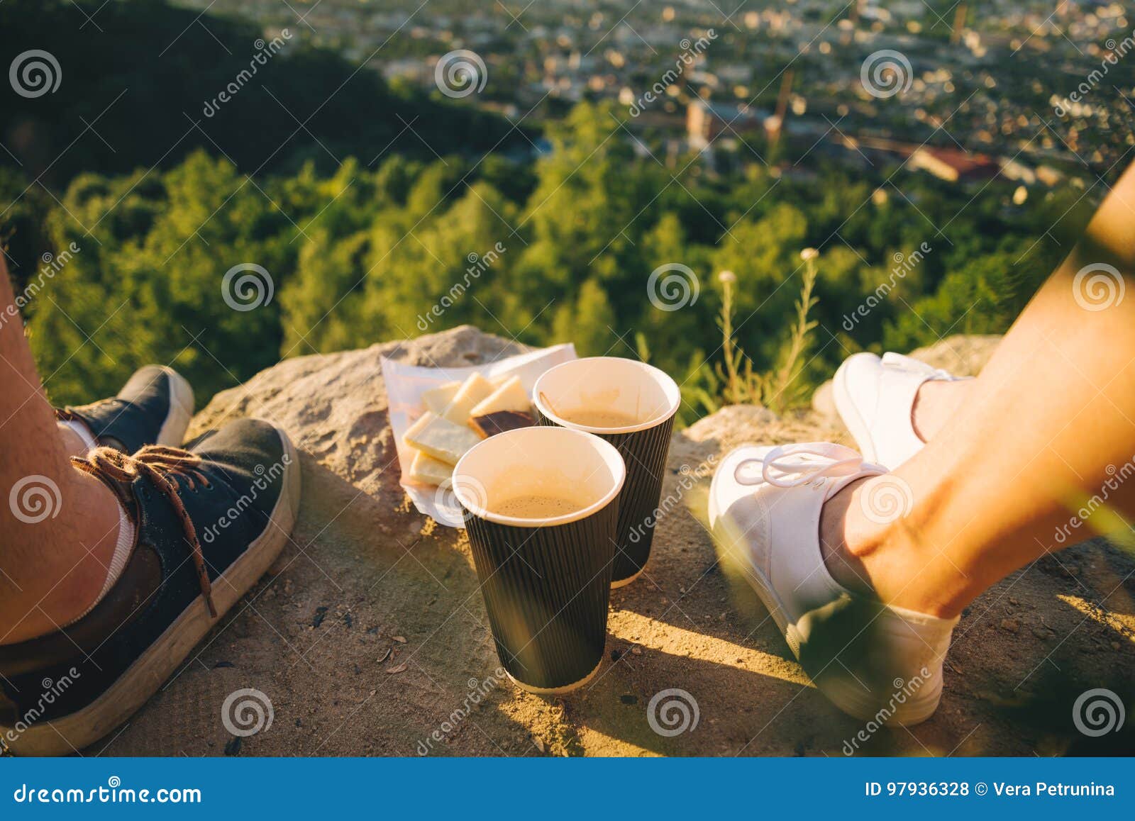 Piernas del hombre y de la mujer en el top de la colina con dos tazas y chocolates, fecha en puesta del sol