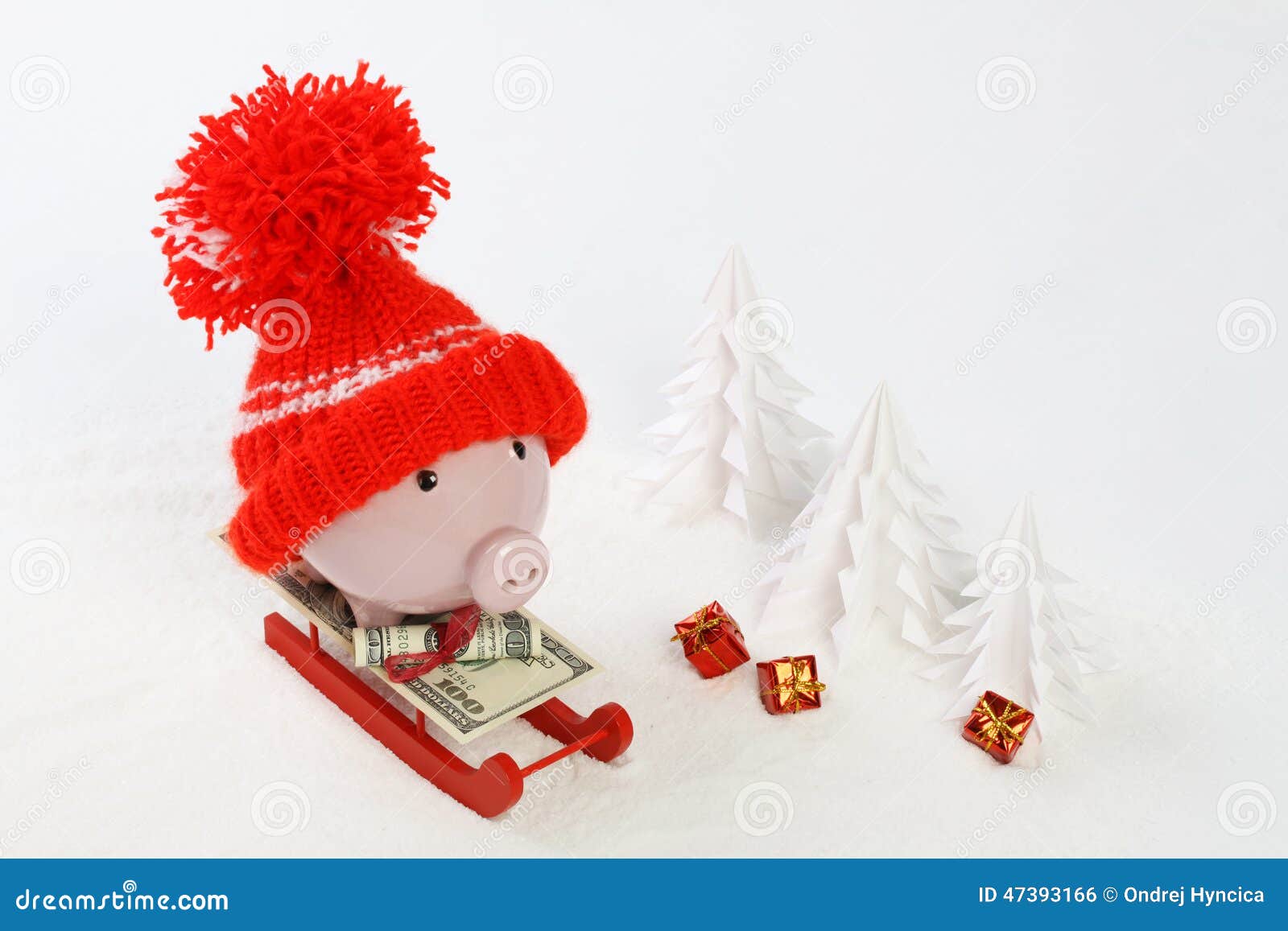piggy box with red hat with pompom standing on red sled with blanket from greenback hunderd dollars on snow and around are