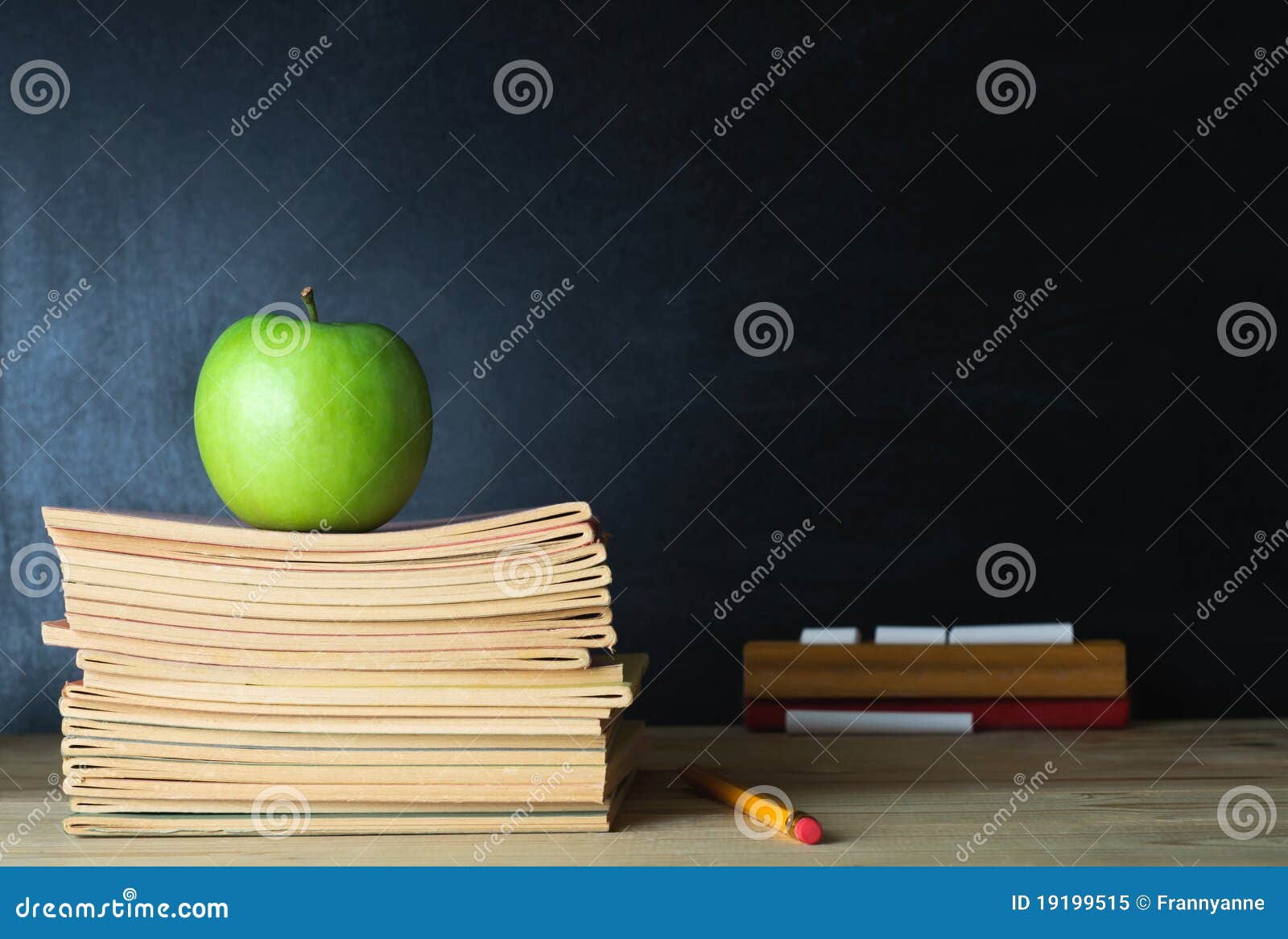 Pizarra de la escuela y escritorio del profesor. Escritorio de un profesor de escuela con la pila de cuadernos y de la manzana en marco izquierdo. Una pizarra en blanco en fondo suave del foco proporciona al espacio de la copia.