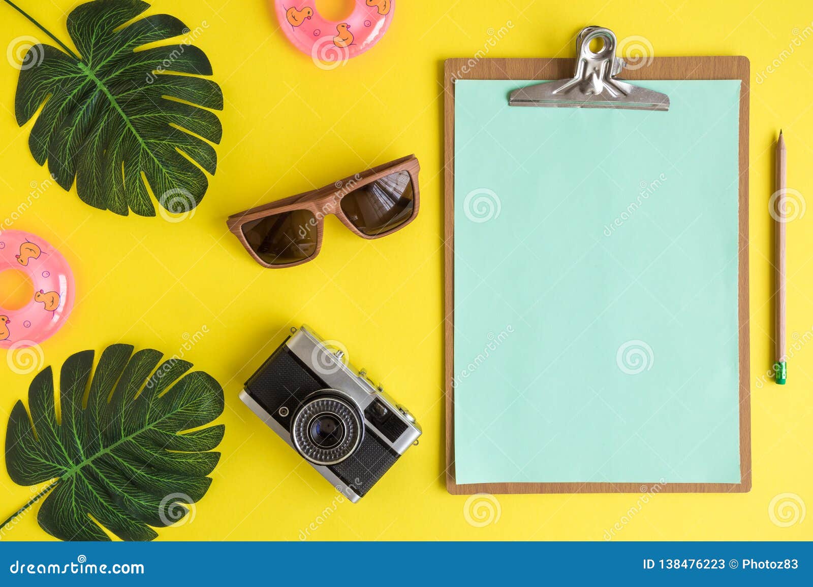 Placa de clipe de papel vazia com óculos de sol, câmera da foto, o flutuador inflável e o sumário do verão das folhas do monstera. Configuração lisa da prancheta com papel vazio com artigos das férias de verão contra o conceito criativo mínimo do fundo amarelo