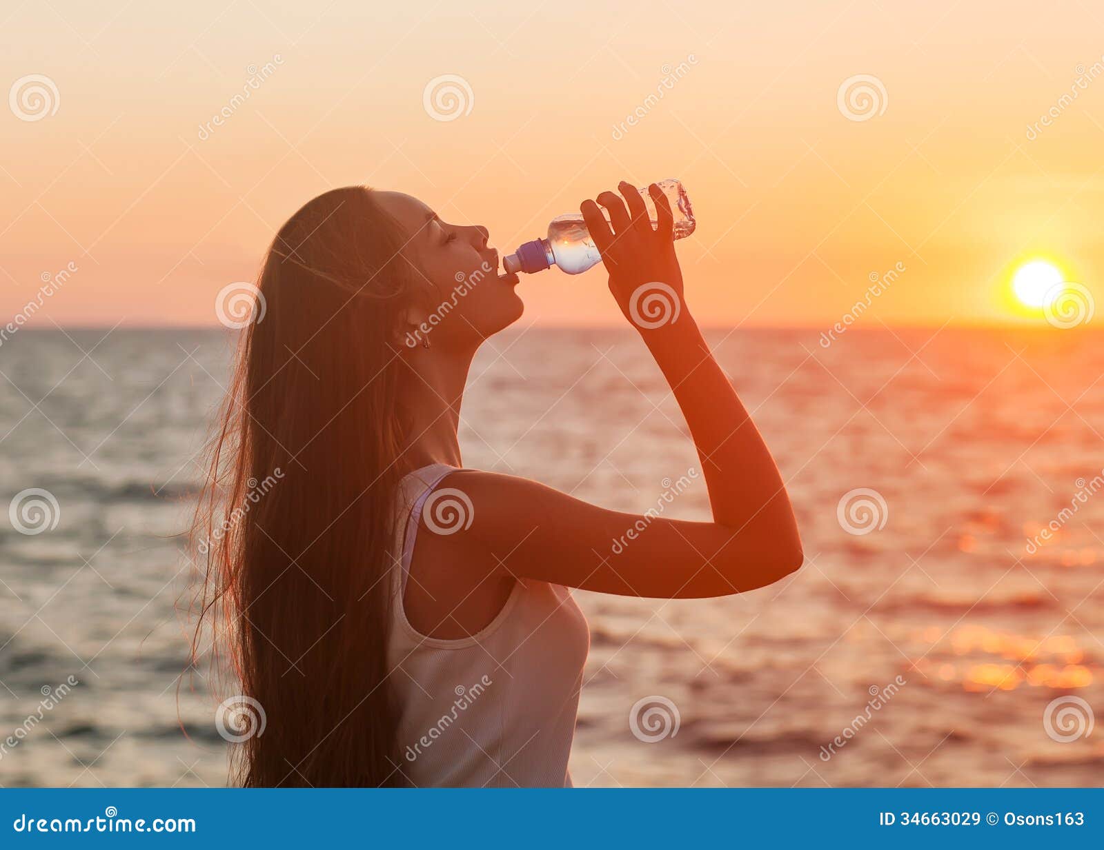 Plaisir - femme heureuse libre appréciant le coucher du soleil. Modèle femelle caucasien asiatique. Femme buvant d'une bouteille d'eau au coucher du soleil