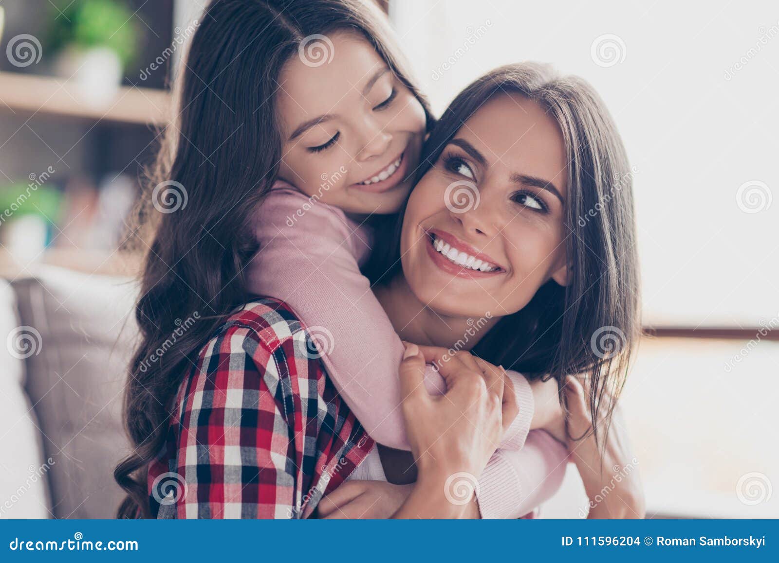 playful small girl with long dark hair is hugging her mum`s neck