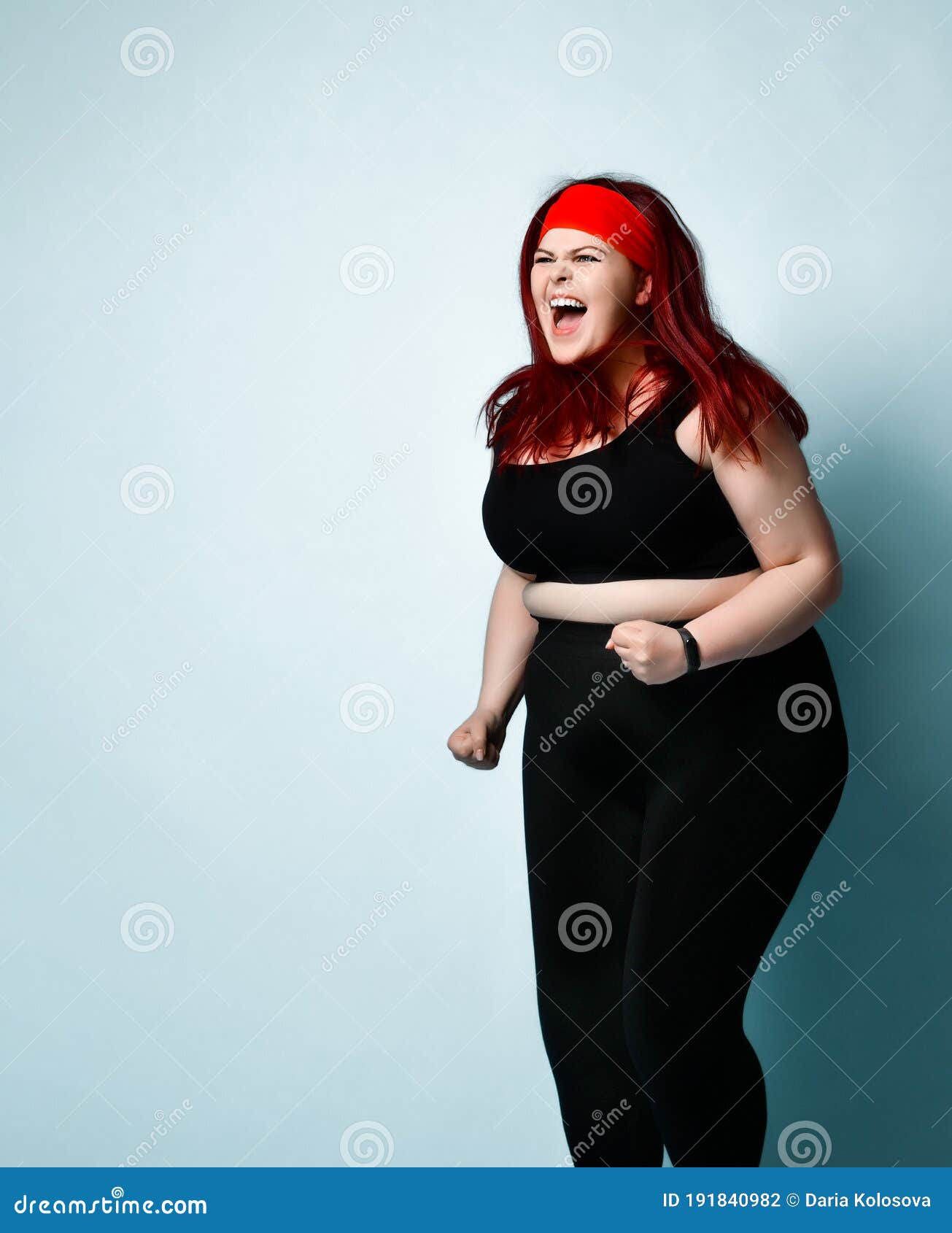 plus-size girl with red hair in black sportswear shouts with anger and expression keeping fists clenched on blue background.