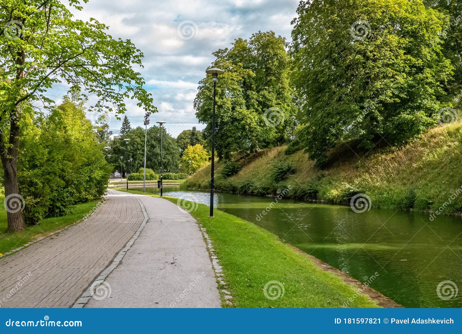 a pond in snelli park, tallinn, estonia. green trees and sidewalk on summer day with clouds