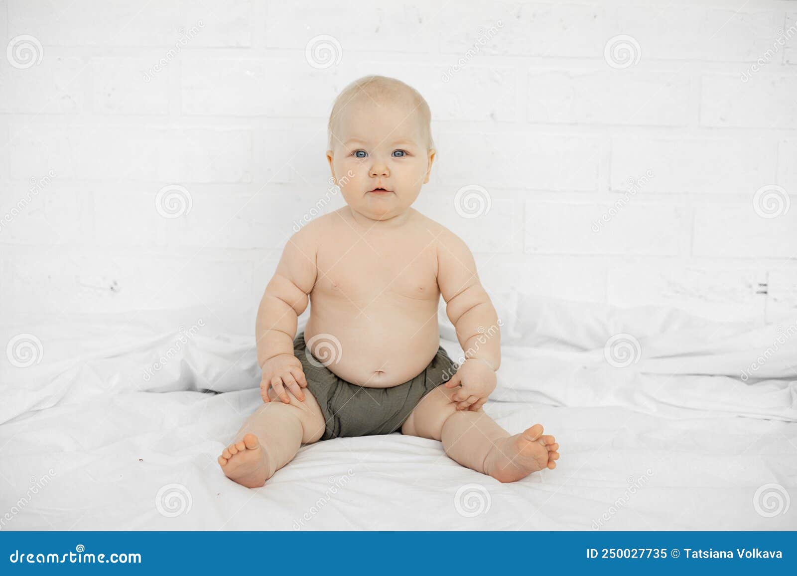 portrait of attractive serious grey-eyed plump cherubic baby infant toddler wearing grey pants sitting on white bed.