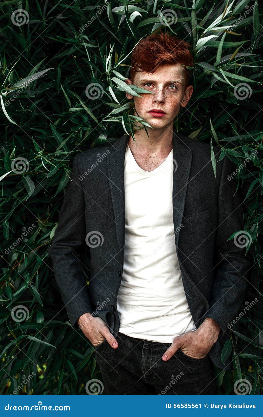 portrait of attractive stylish young guy model with red hair and freckles sitting on green grass, wearing jacket. fashionable outd