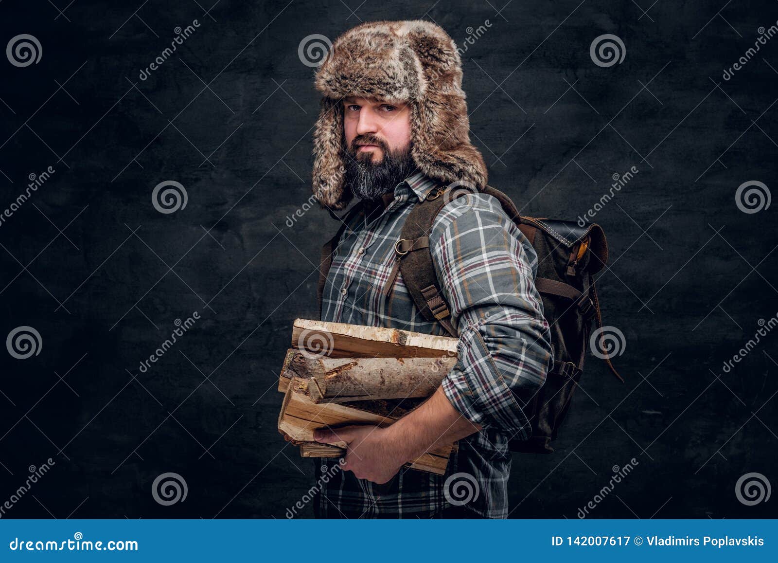 portrait of a bearded woodcutter with a backpack dressed in a plaid shirt and trapper hat holding firewood.