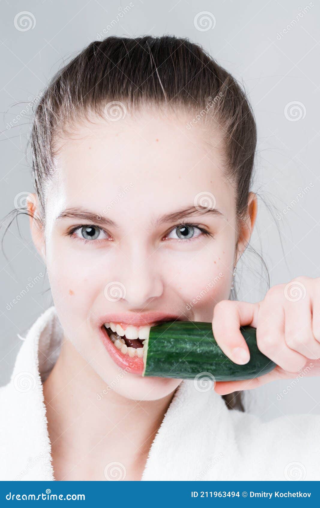 Portrait Of A Beautiful Woman Holding A Cucumber And Biting It On A Gray Ba...