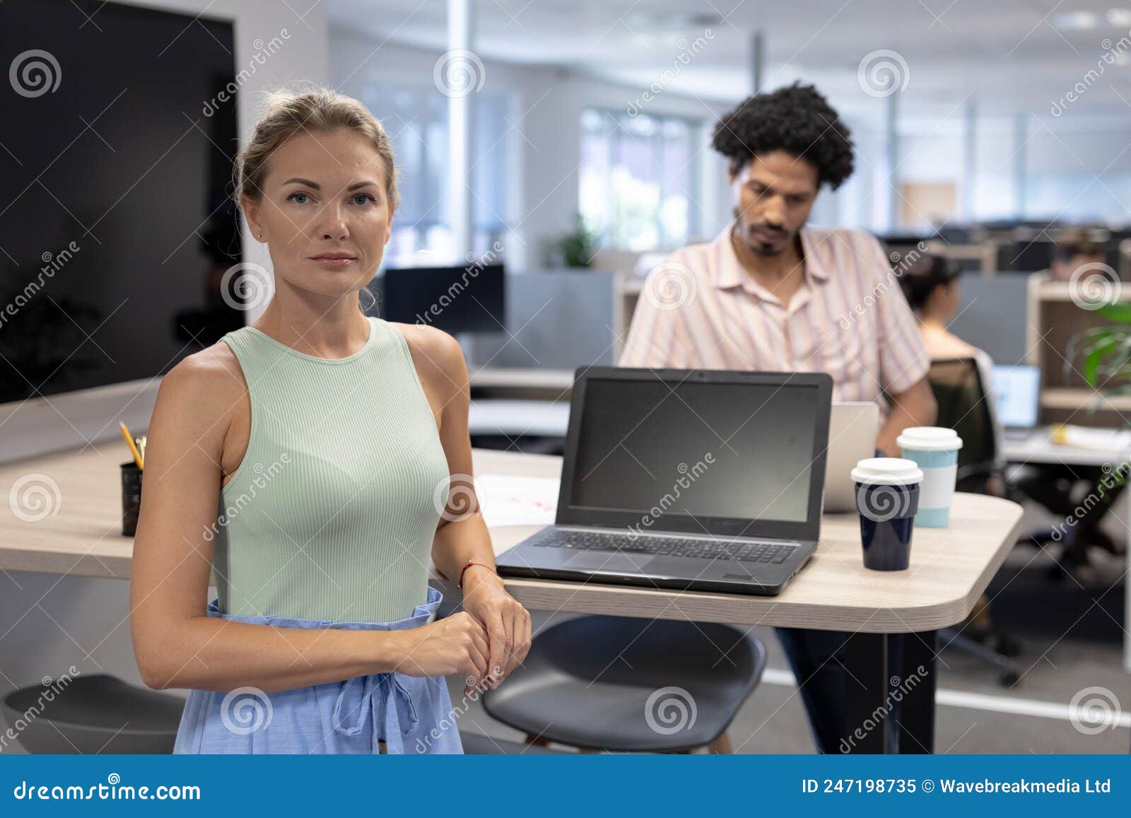 portrait of confident cauasian businesswoman standing by laptop against biracial businesswoman