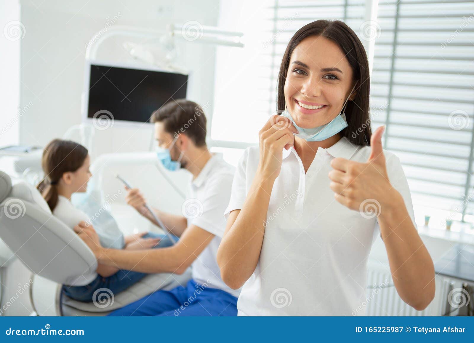 portrait of female dentist showing thumb up while her collegue working with small client on the background