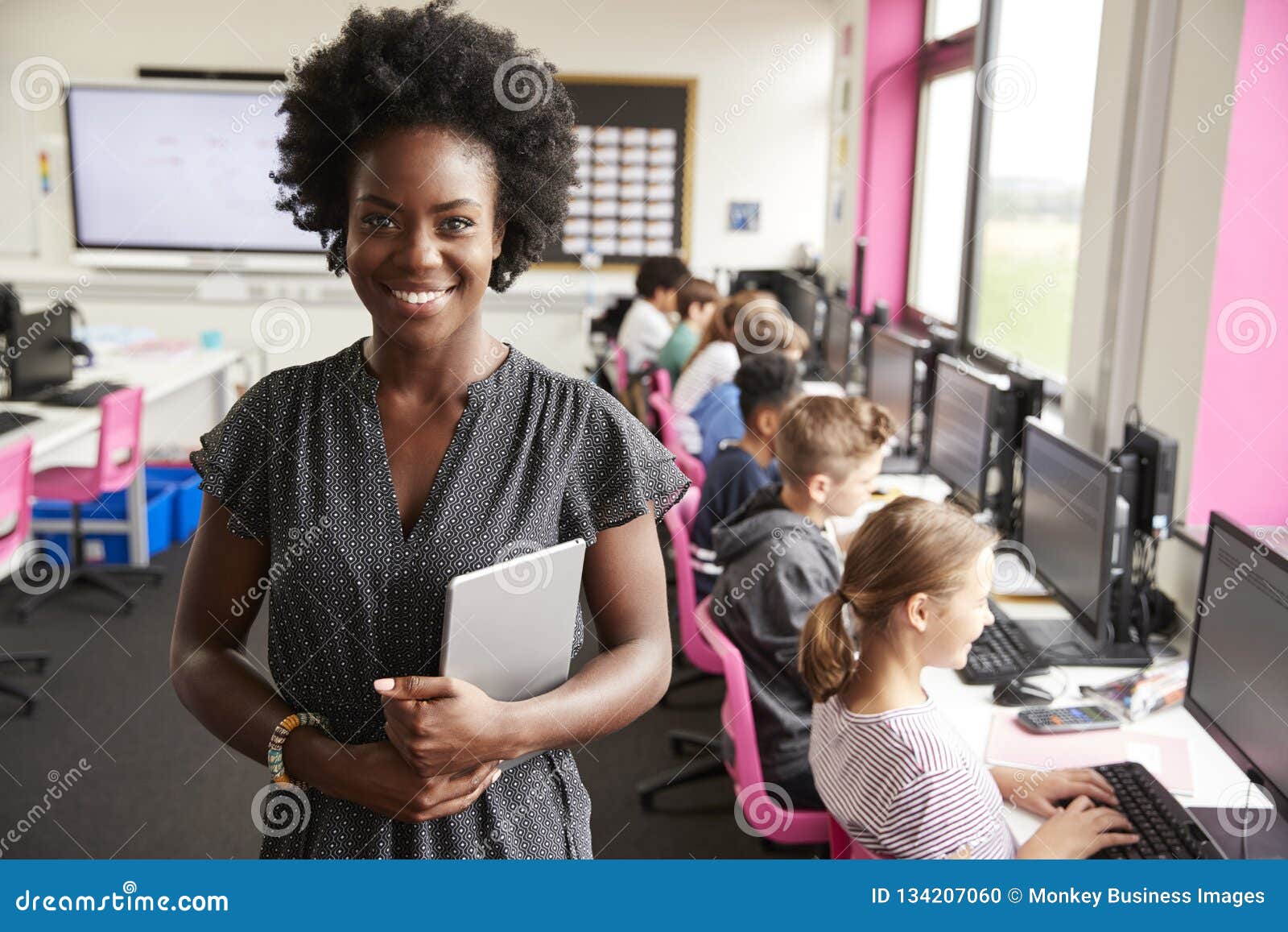 portrait of female teacher holding digital tablet teaching line of high school students sitting by screens in computer class