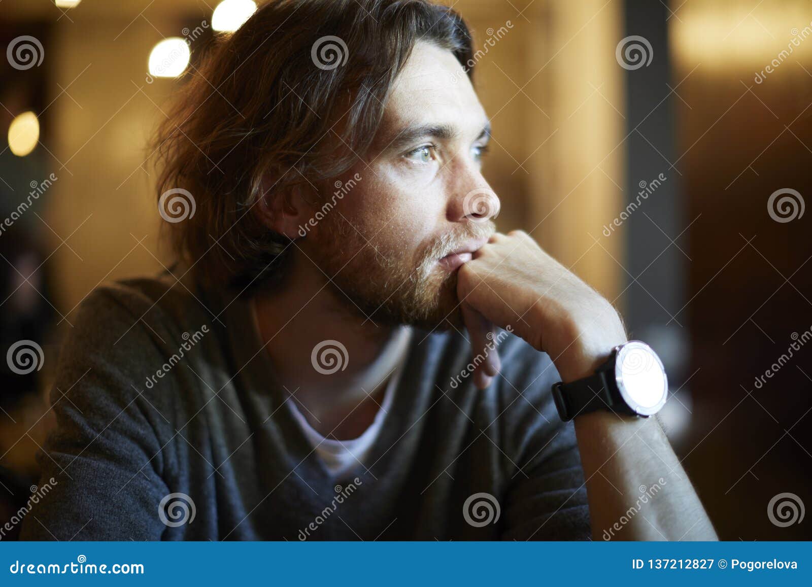 portrait of handsome hipster guy with long hair and beard sitting in sunny cafe, resting near window. romantic man looks lonely.