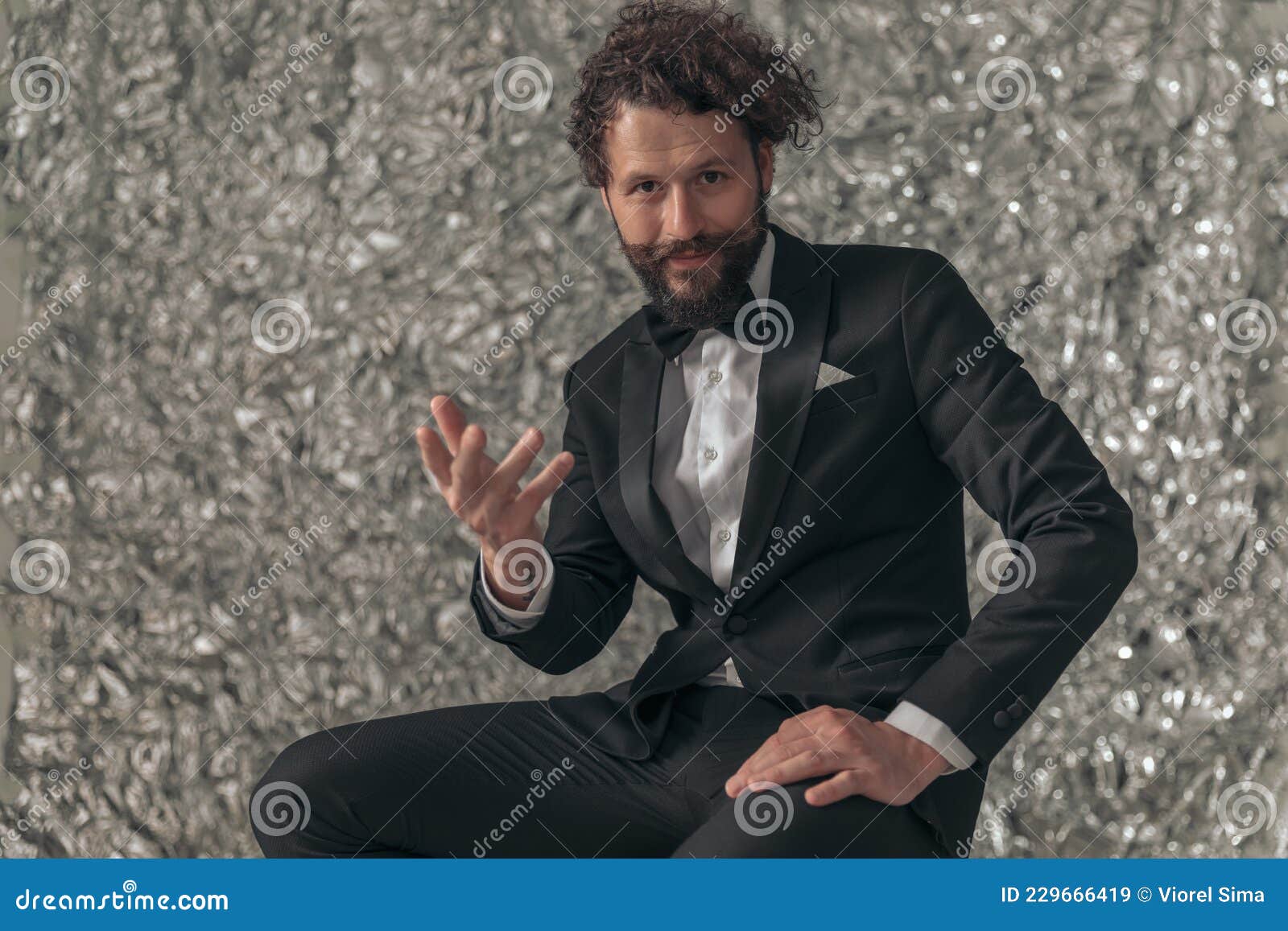 happy bearded elegant man in black tuxedo holding hand up and gesticulating