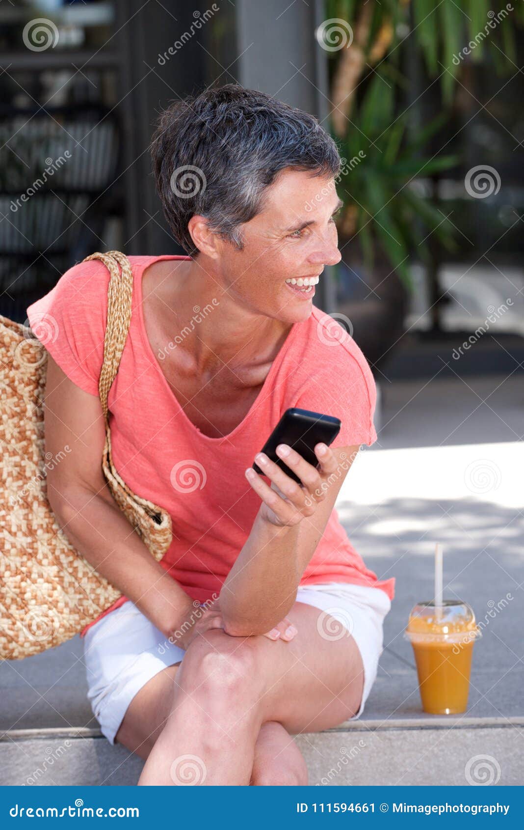 Happy Mature Woman Sitting Outdoors With Drink And Using Cellphone