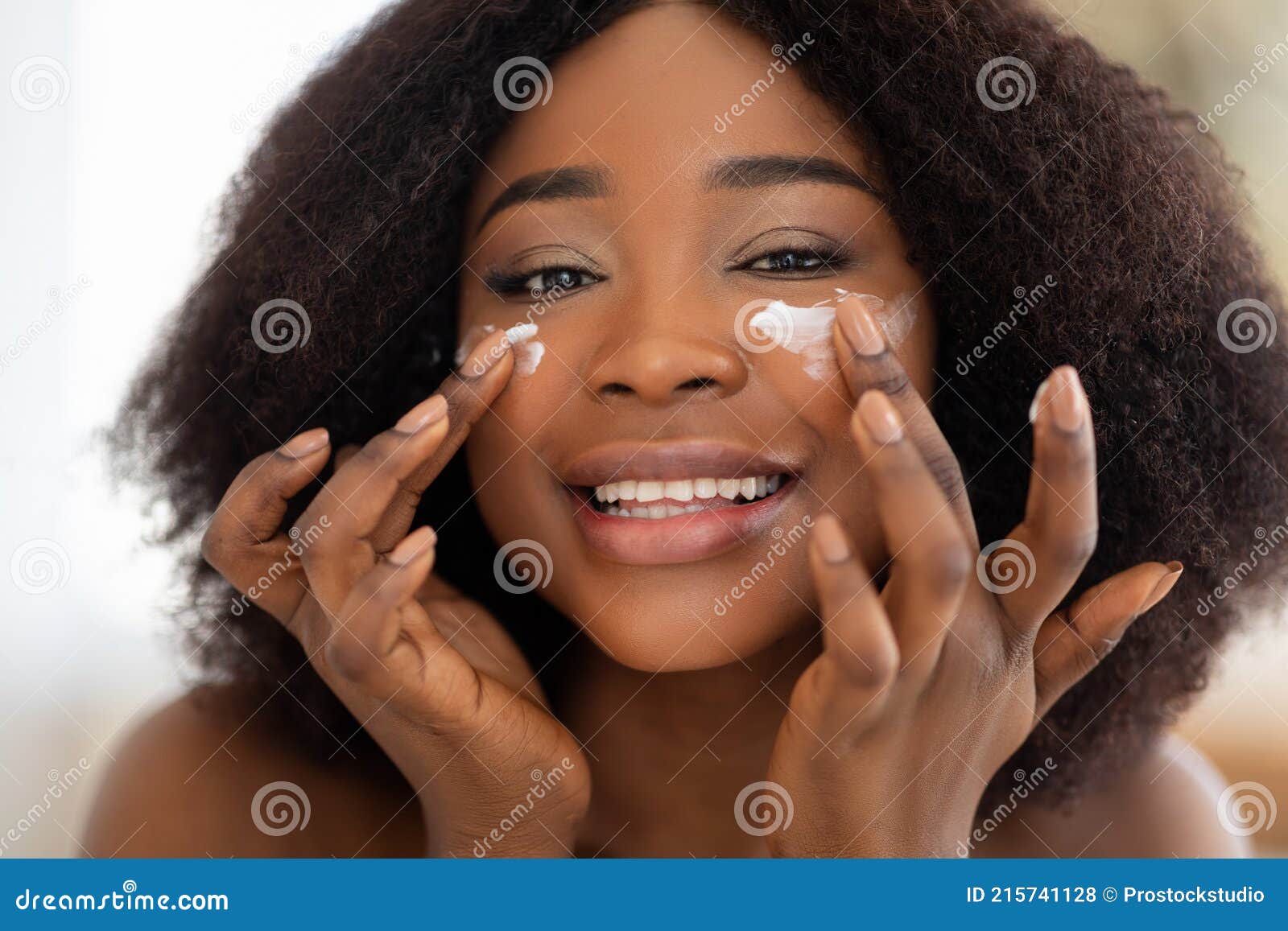 portrait of lovely black woman applying face cream onto her cheeks, smiling at camera, indoors