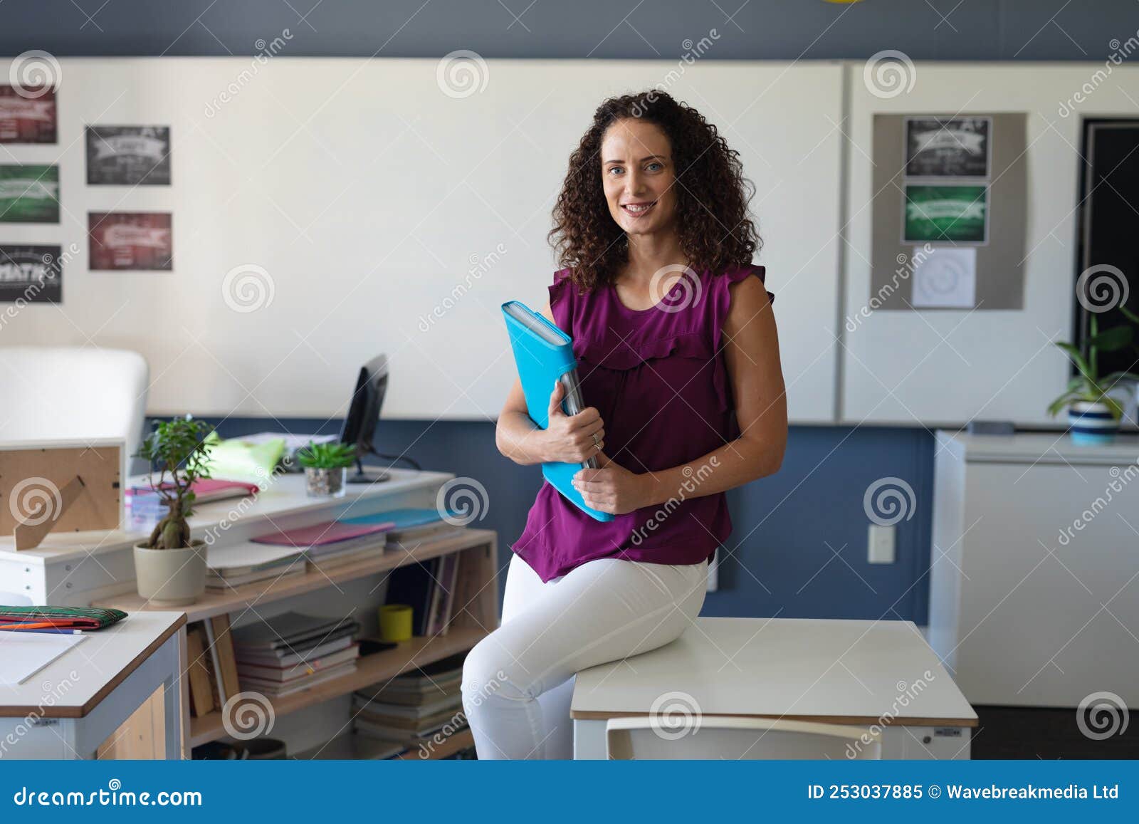 portrait of smiling caucasian young female teacher with folder sitting on table in staffroom