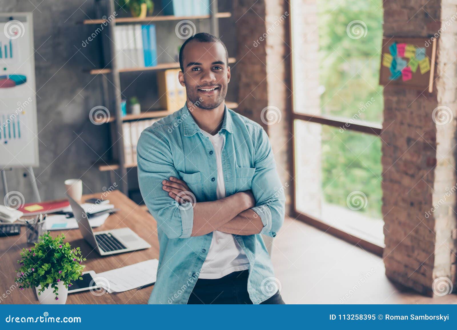 portrait of successful black guy, looking at the camera, standing with crossed arms at his work place in the office, in casual sma