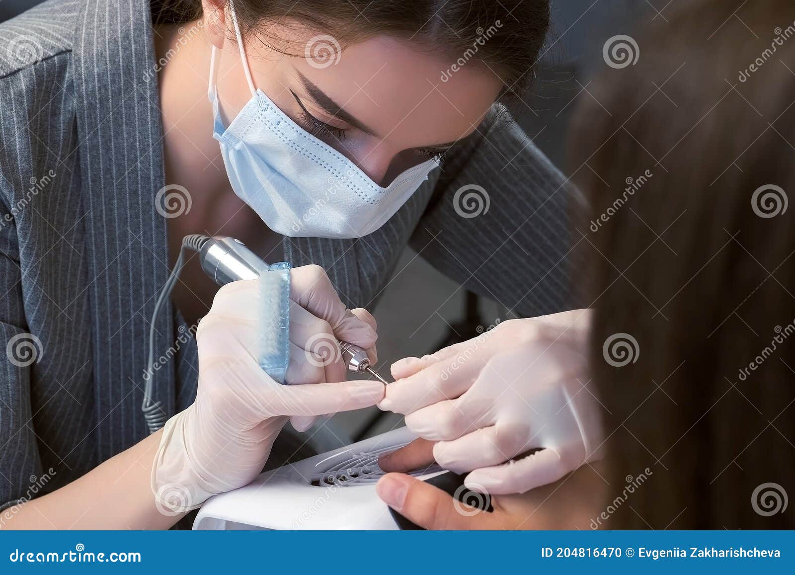 portrait of woman manicurist in mask cleaning pterygium on nail using apparatus.