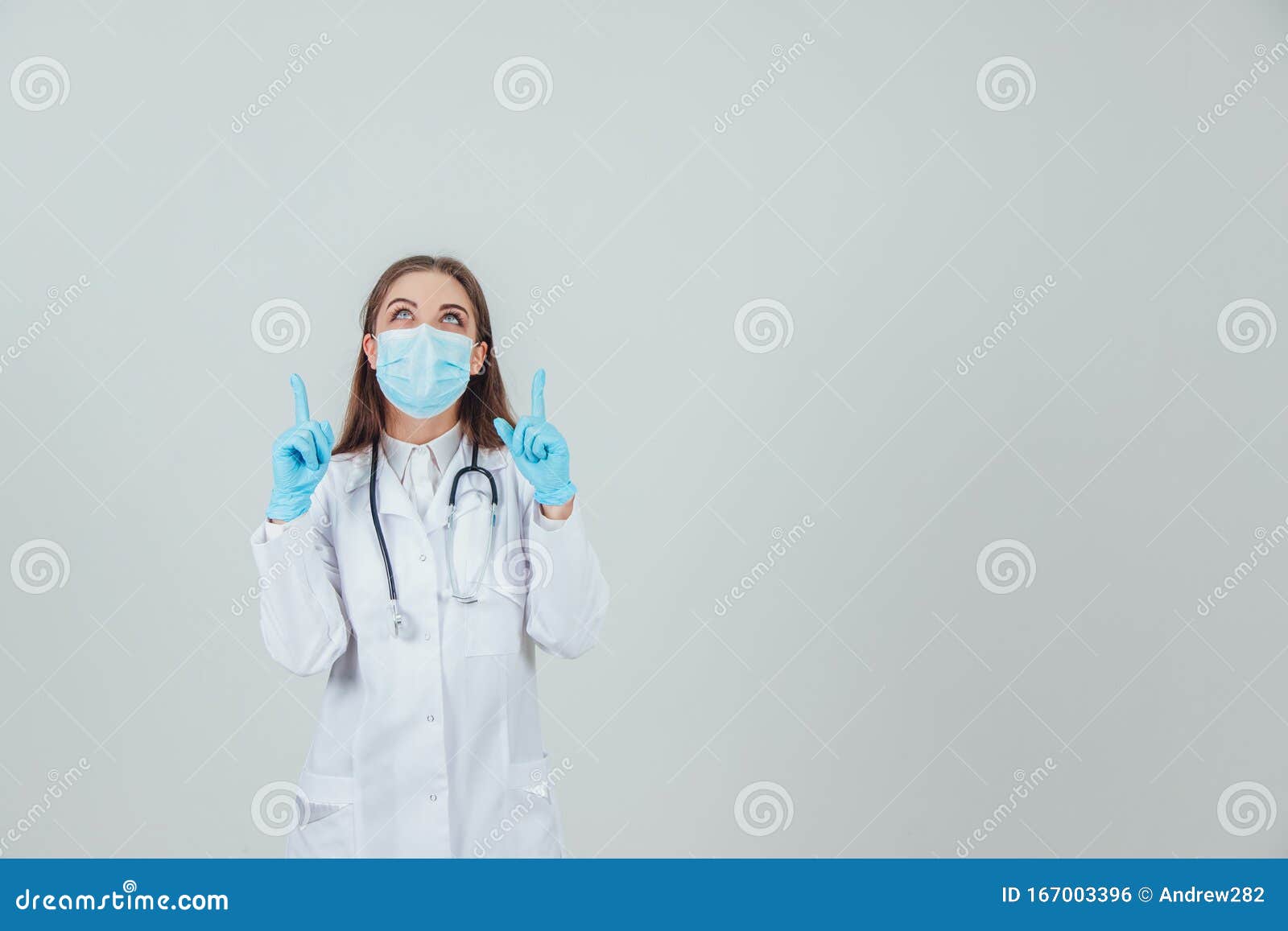 portrait of a young doctor in medical mask, on sterile gloves and with stethoscope, pointing her fingers up.