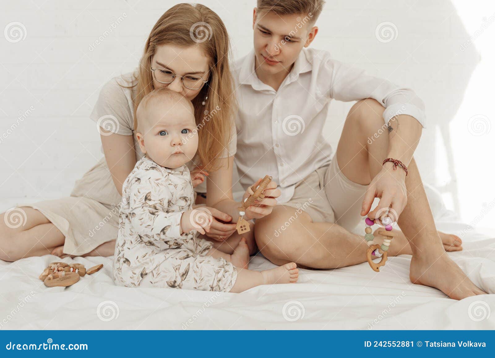 portrait of young family with cherubic infant baby, with blue eyes holding wooden natural teether sitting on white bed.