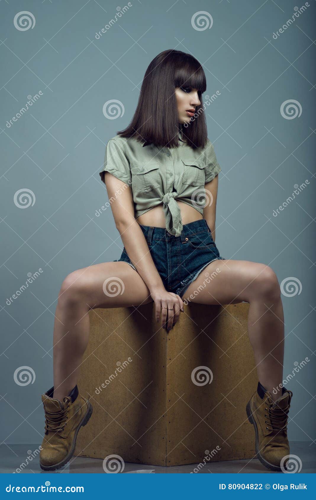 portrait of young gorgeous dark-haired model wearing high waisted dark blue jeans shorts, khaki shirt and boots sitting on cube