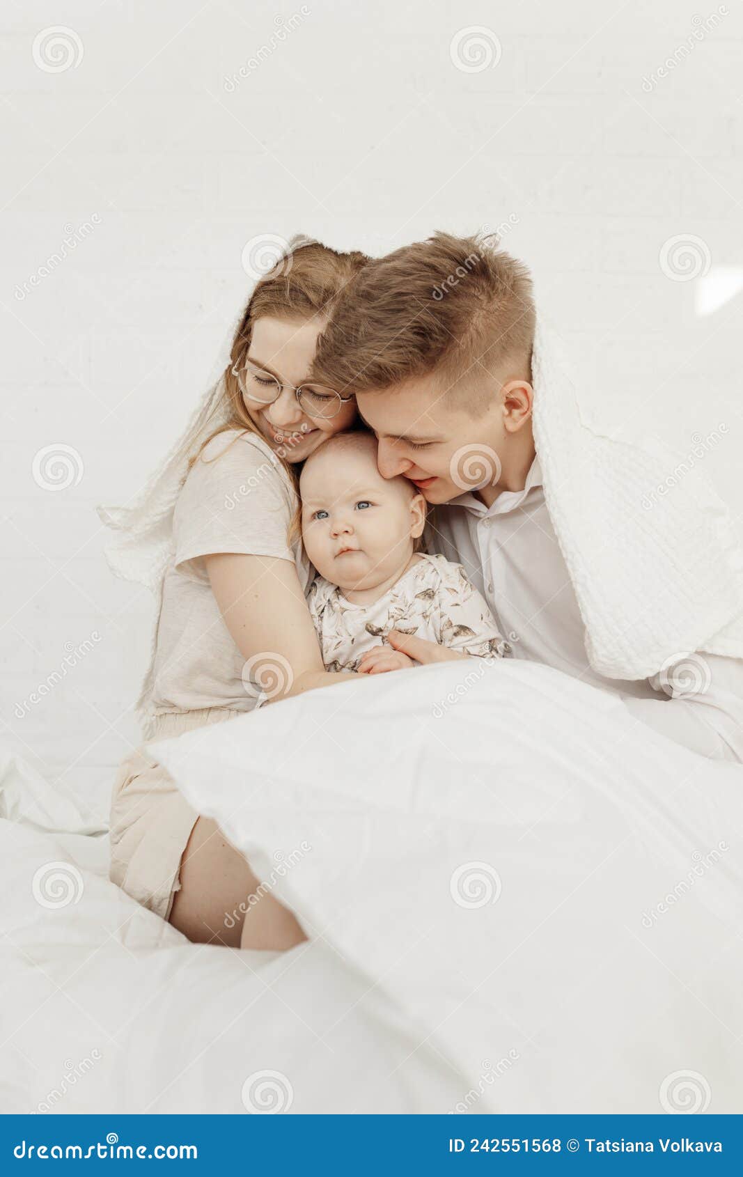 portrait of young happy smiling beautiful family with cute cherubic infant baby in white clothes sitting on white bed.