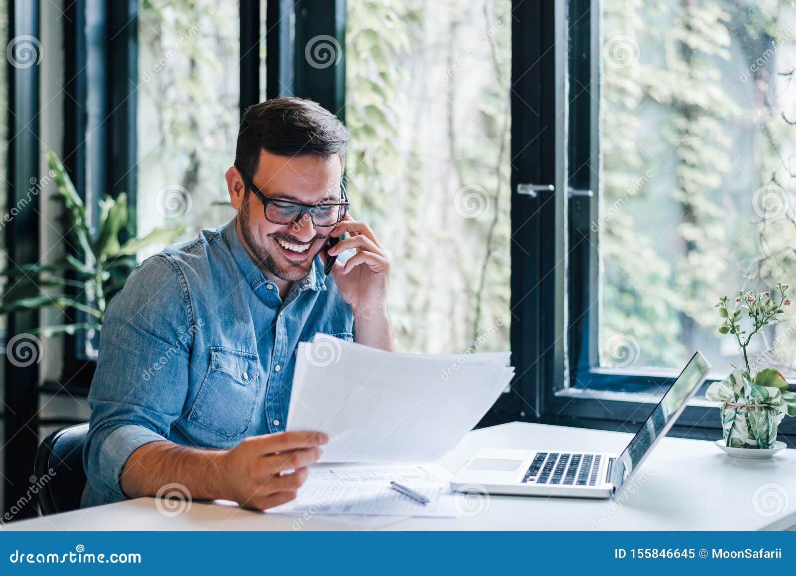 portrait of young smiling cheerful entrepreneur in casual office making phone call while working with charts and graphs looking at