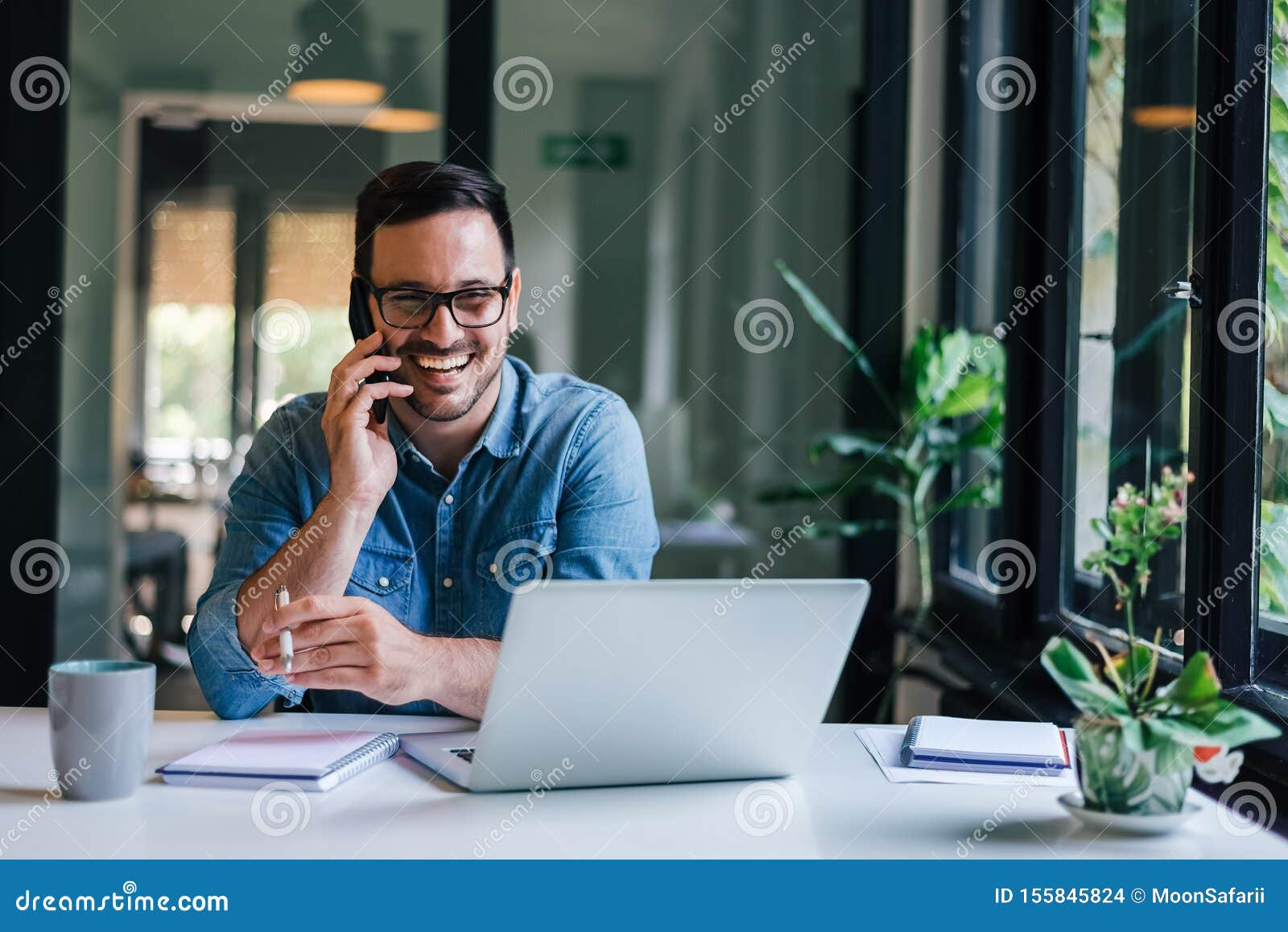 portrait of young smiling cheerful entrepreneur in casual office making phone call while working with laptop