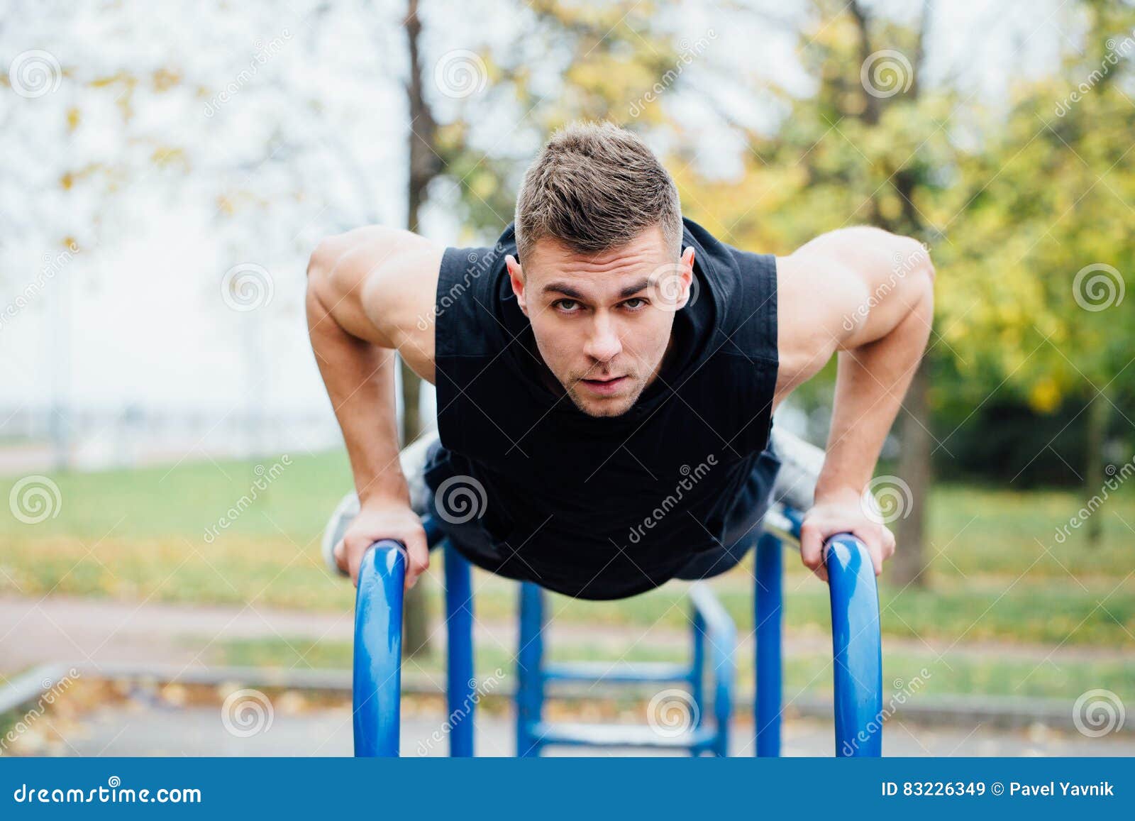 Portret van de geconcentreerde spier jonge mens in zwarte trainingkleren die onderdompelingen op brug doen. Een portret van een geconcentreerde spier jonge mens in zwarte trainingkleren die onderdompelingen op brug doen bij het parkochtend van de dalingsstad