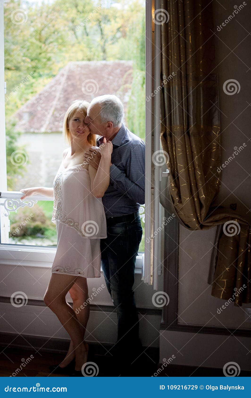 Portret van Sexy Vrouw die Op middelbare leeftijd van Omhelzing van Haar Bejaarde Echtgenoot genieten die zich dichtbij Geopend Venster binnen Hun Huis bevinden Paar met leeftijdsverschil