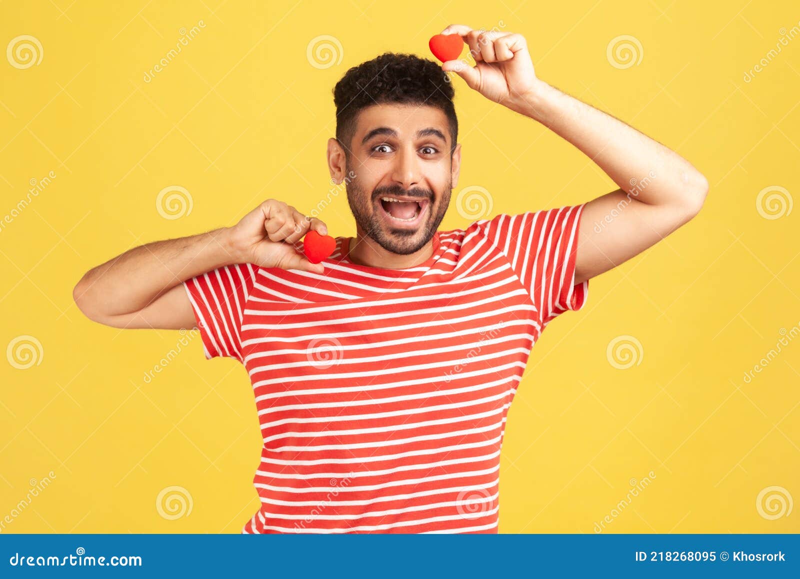 positive excited bearded man in striped t-shirt fooling around having fun with red toy hearts, showing his fondness and devotion,