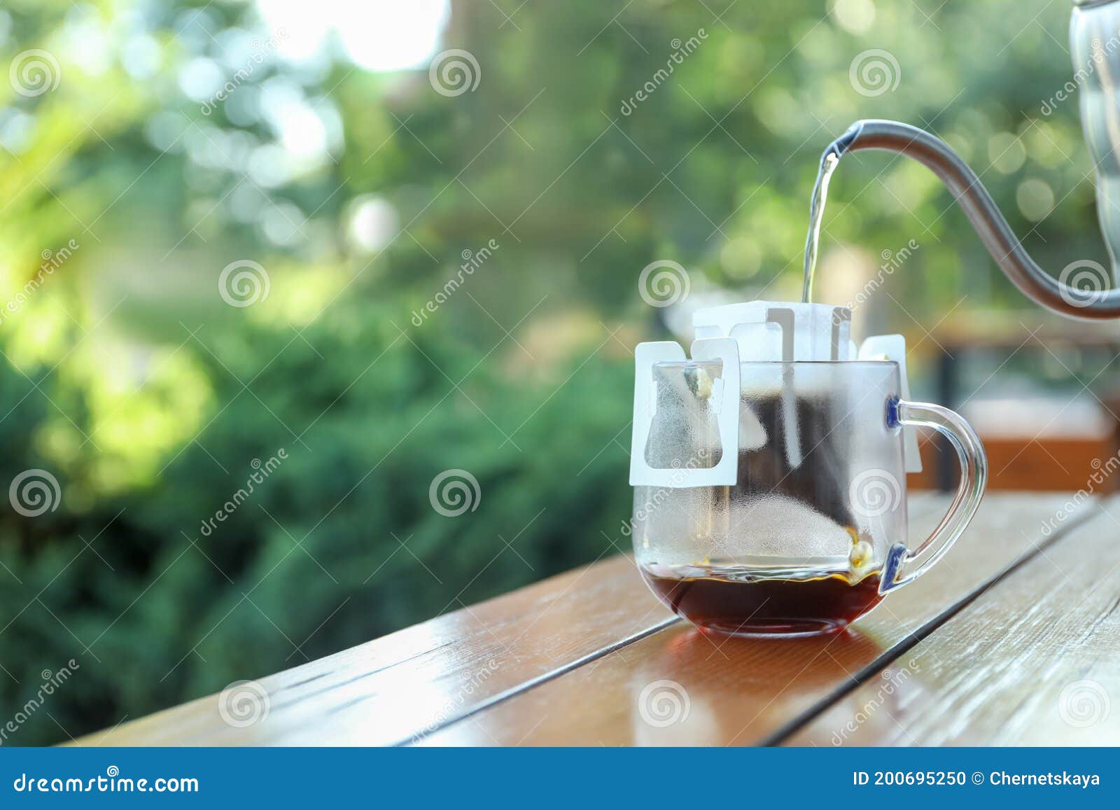 Pouring Hot Water Into Glass Cup With Drip Coffee Bag From Kettle On Wooden Table Outdoors