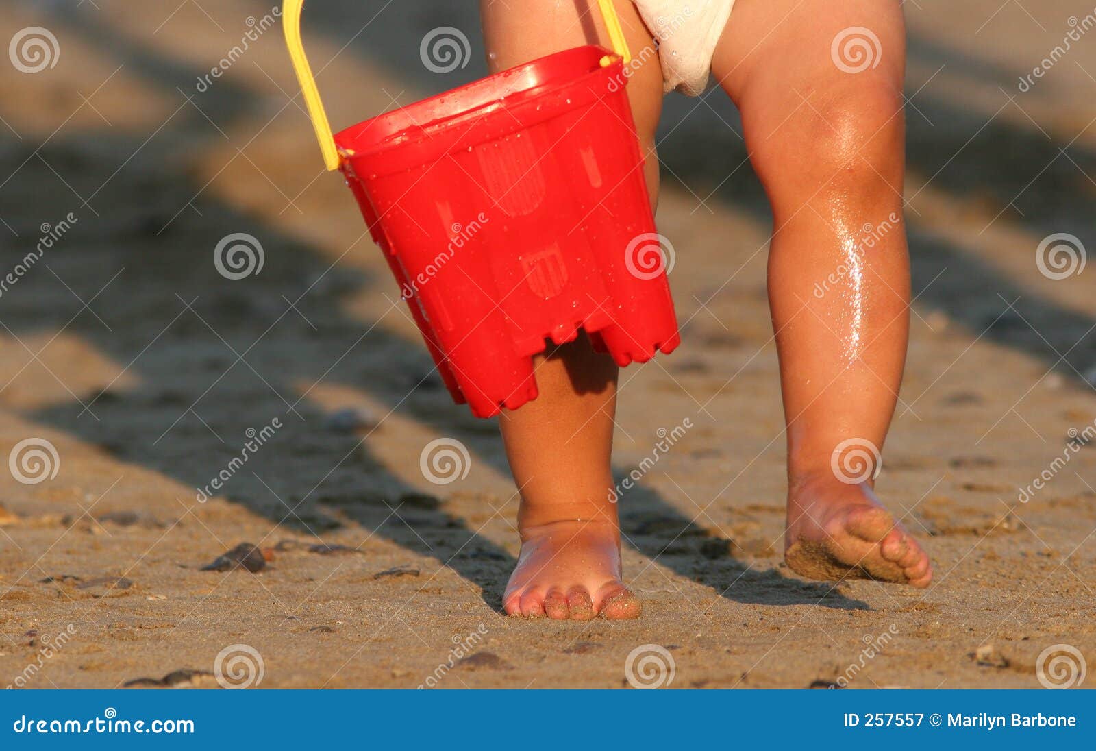 Prazeres primordiais. Os pés molhados e os pés arenosos de uma criança que desgasta uma fralda e que carreg uma cubeta plástica vermelha que anda em uma praia.