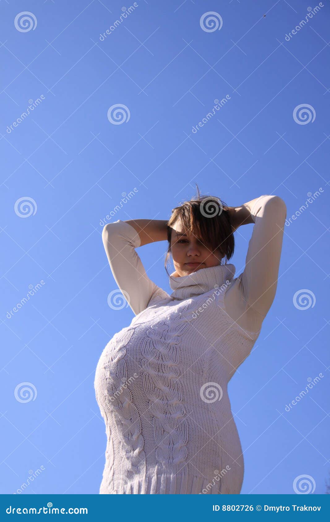 Pregnant girl against the sun. Pregnant girl in a white sweater with high collar holding hands behind the head on a blue sky background against the sun