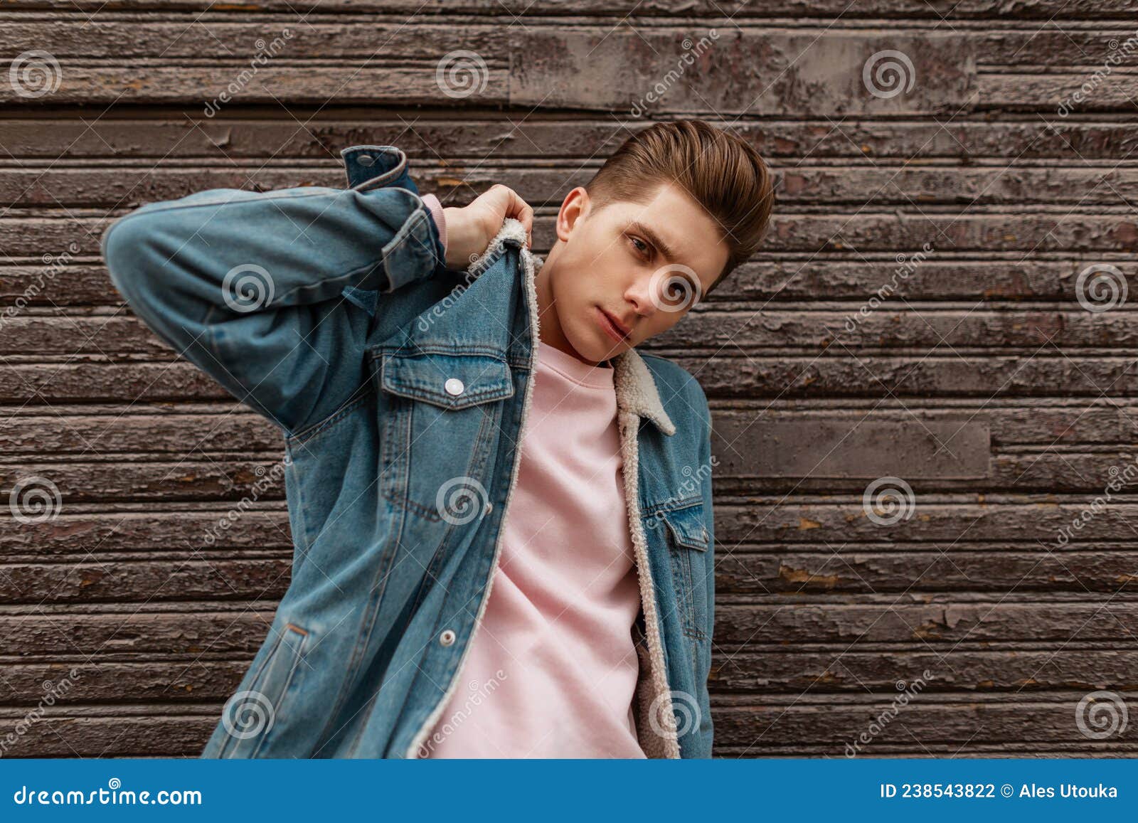 Model Of A Fashionable Young Blonde Woman In Stylish Blue Jeans In A Trendy  Shirt With A Hairstyle Stands Near A White Wall In The Room. Beautiful  Elegant Girl In Youth Casual