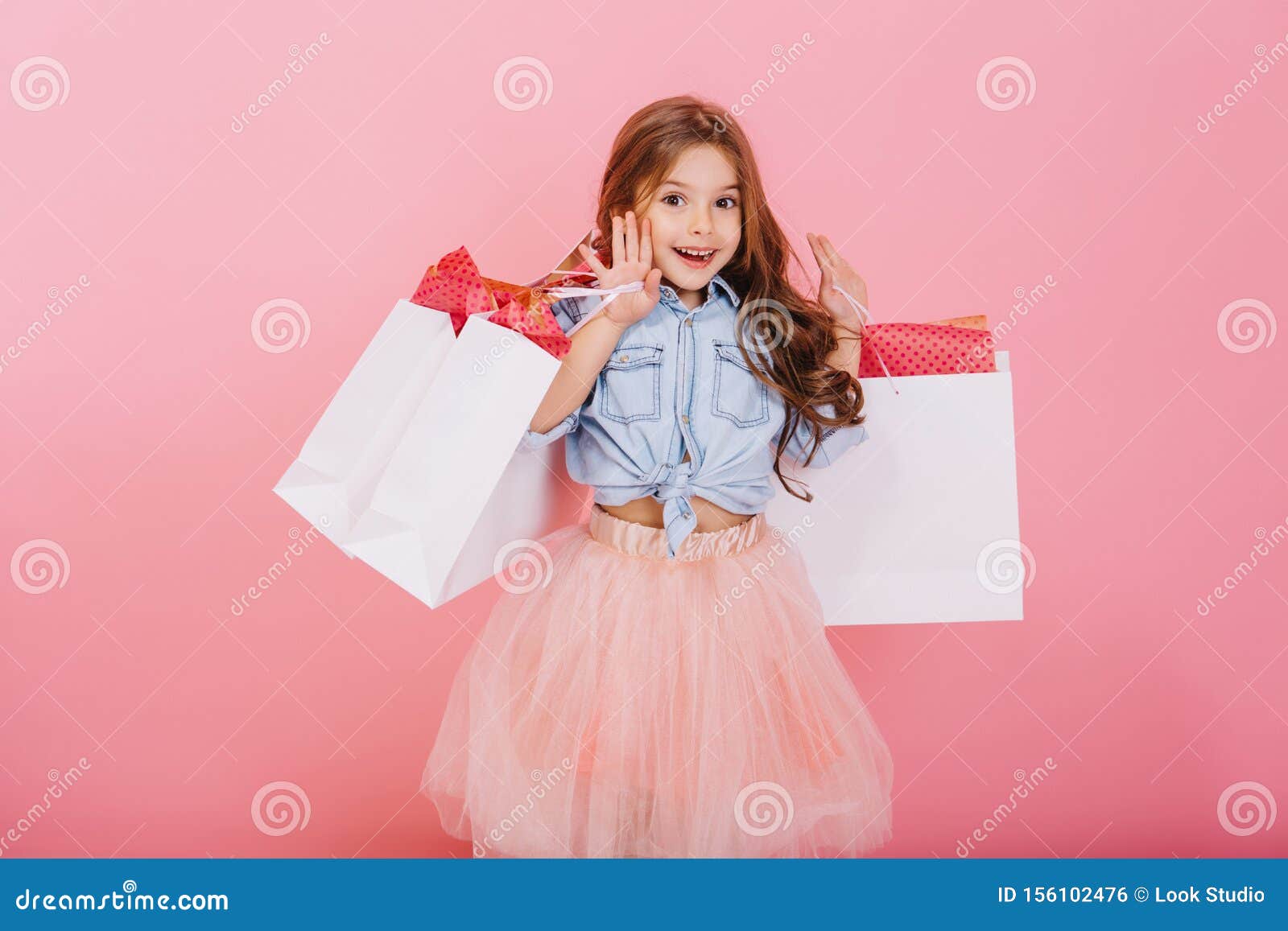 pretty joyful young girl in tulle skirt, with long brunette hair walking with white packages on pink background. lovely